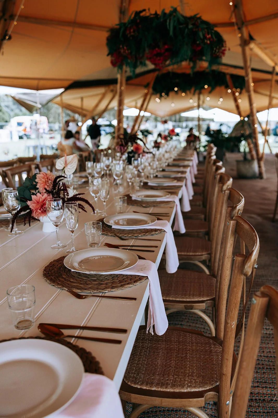 Long table set for a wedding reception under a tent, with wooden chairs, plates, glasses, cutlery, pink napkins, and floral centerpieces.