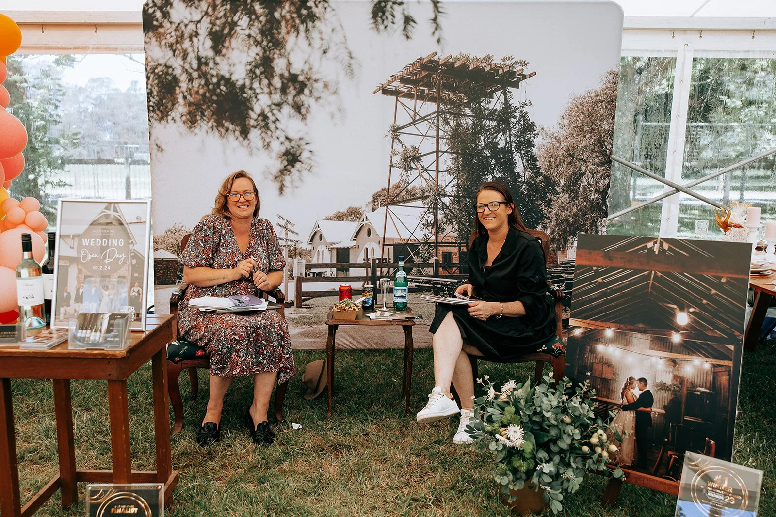 Two women seated outdoors at a wedding open day booth, surrounded by large wedding-themed photos and décor, with informational material and drinks on tables.