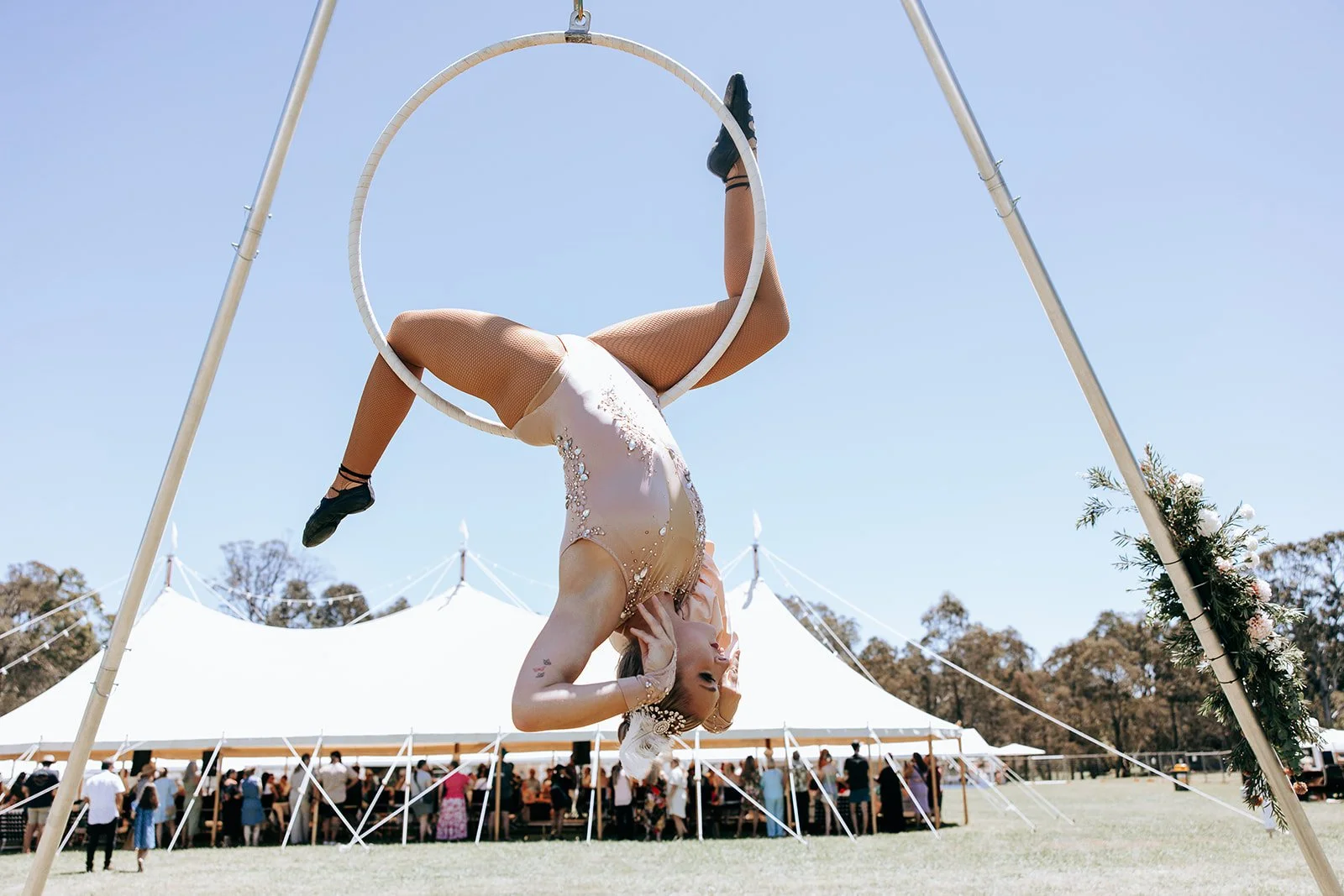 Aerial performer on hoop under a large tent outdoors with audience watching.