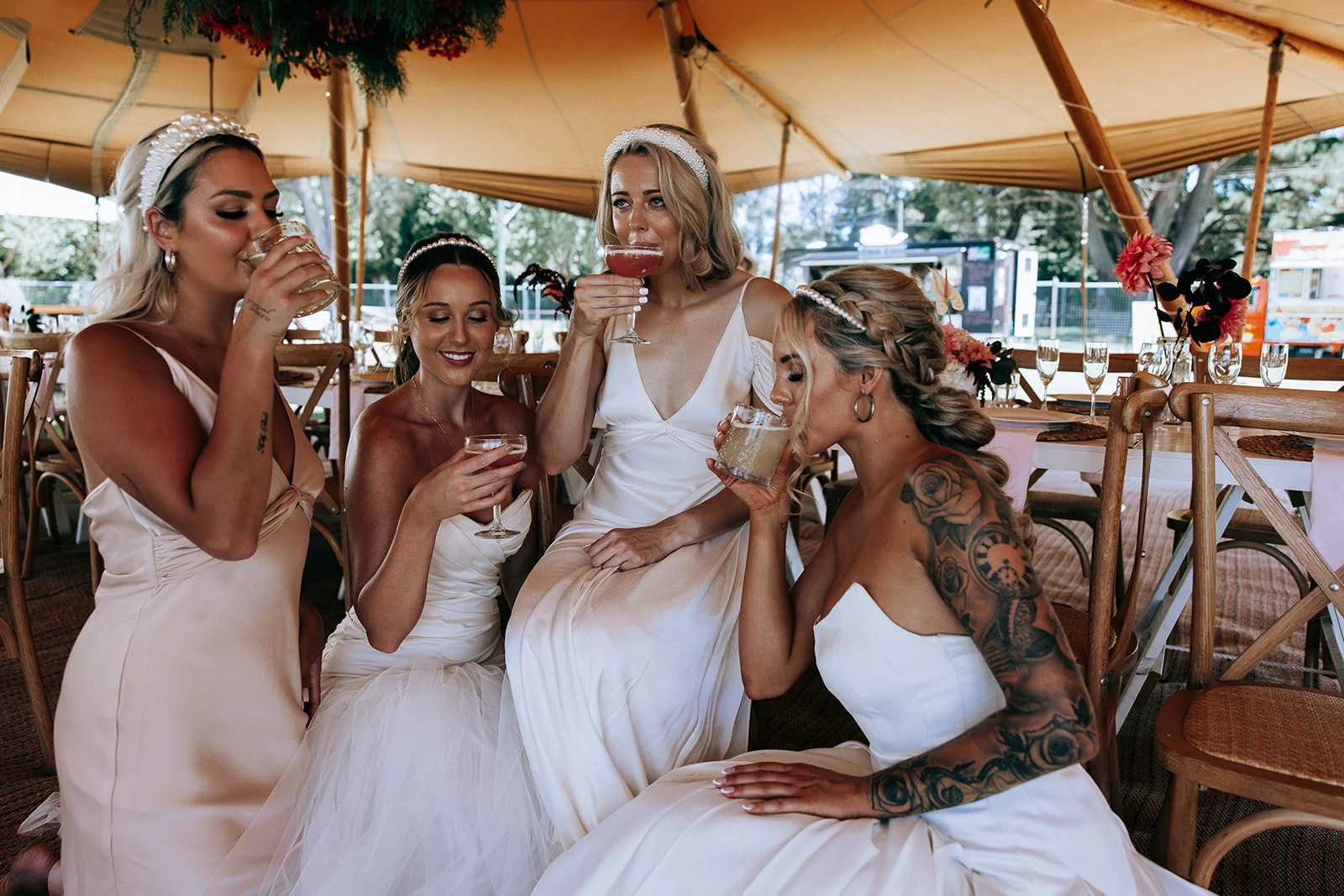 Four women in elegant dresses drinking from glasses under a tent at an outdoor event.