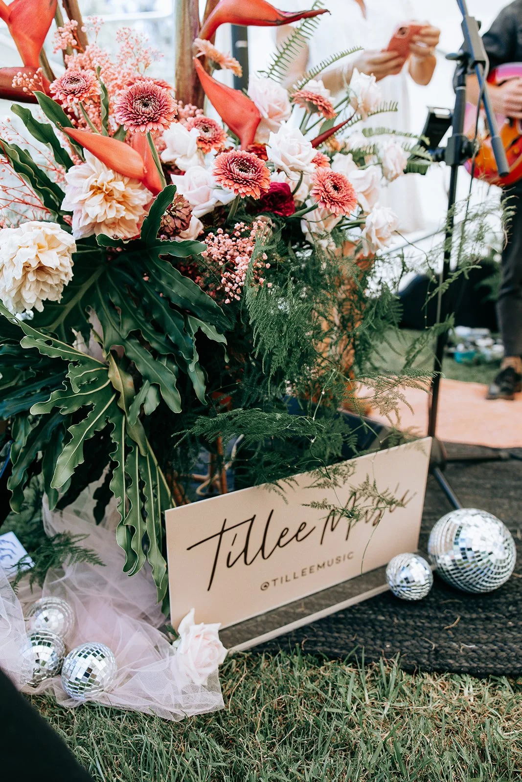 Colorful floral arrangement with calla lilies, gerbera daisies, roses, and greenery, surrounded by small disco balls and a sign reading "Tillee Music" on grass.