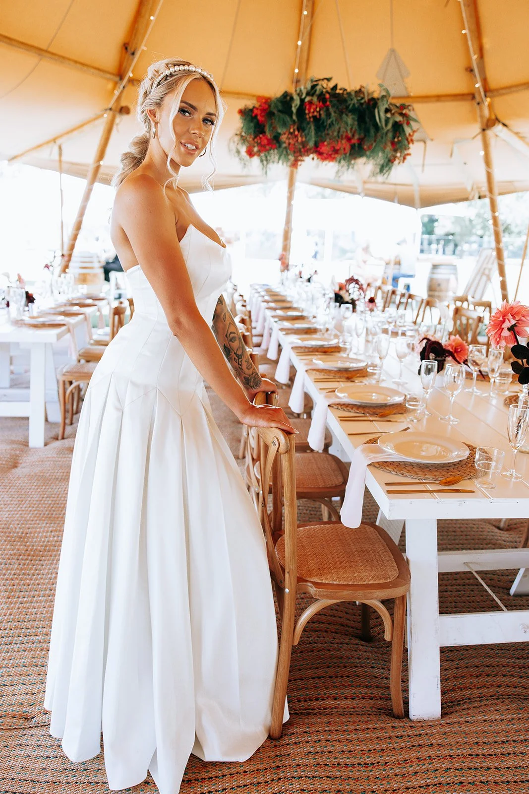 Bride in a white wedding dress stands beside a long, elegantly set dining table under a large tent. The table is decorated with plates, glassware, and flowers. Overhead, a floral arrangement hangs from the tent ceiling.
