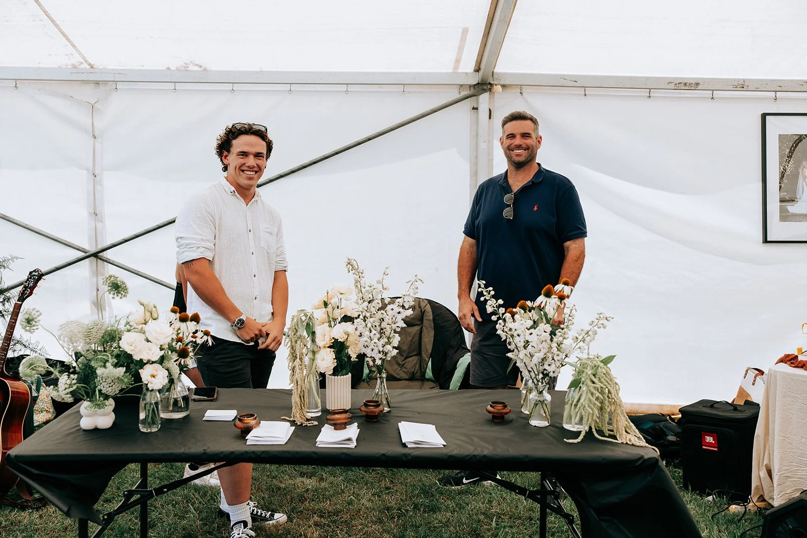 Two men smiling behind a table with floral arrangements in glass vases, inside a white tent. A guitar is visible on the left.