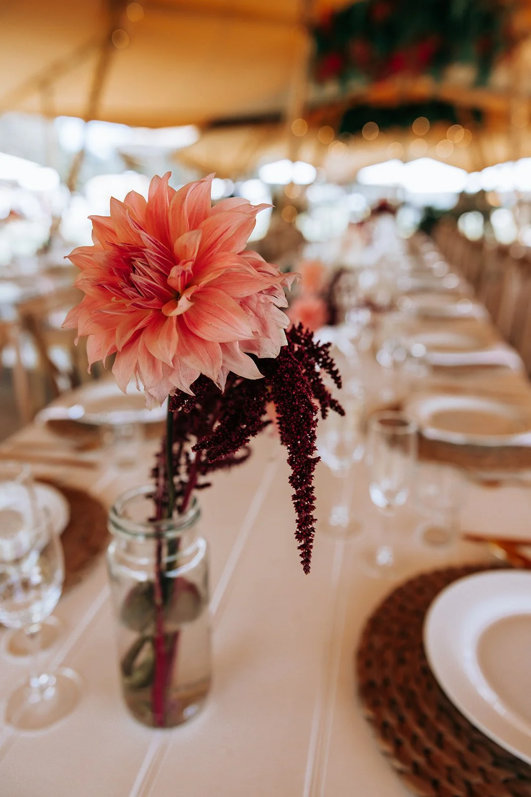 Close-up of a pink flower in a glass vase on a decorated dining table with white plates and woven placemats.