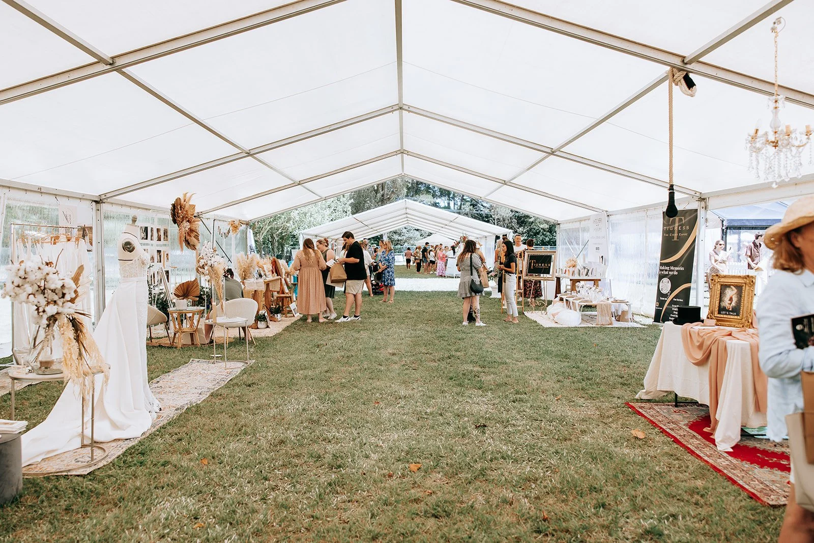 Outdoor market event under a large white tent, featuring various booths with clothing displays and decorative items on grassy area, with people browsing and socializing.