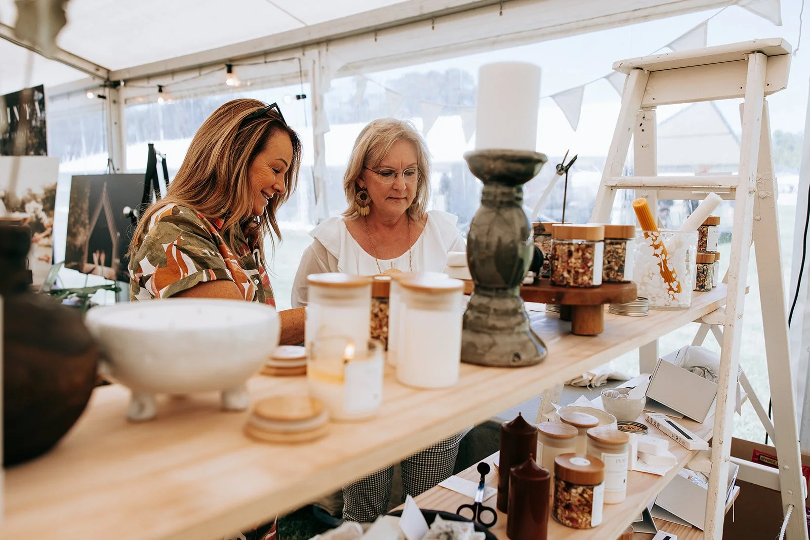Two women at a market stall with candles and home decor, smiling and interacting inside a tent.