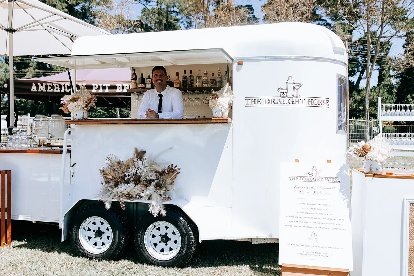 Mobile bar trailer "The Draught Horse" with a server in a white shirt and black tie, featuring a decorative arrangement of dried flowers. Assorted bottles are displayed in the background.