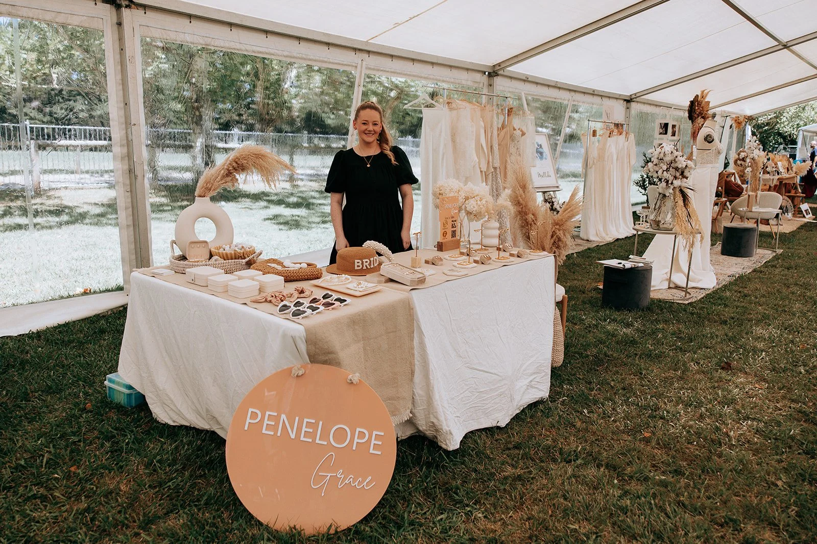 A woman stands behind a table displaying bridal accessories and decor under a tent at a wedding fair. Items include jewelry, a hat labeled 'BRIDE', and decorative pieces. The table is covered with a beige cloth and a sign reading 'Penelope Grace' is 