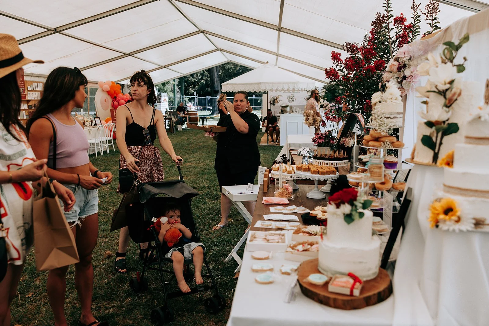 People at an outdoor event under a tent, with a dessert table featuring cakes and pastries. A woman is pushing a stroller with a child, and others are browsing the food offerings.
