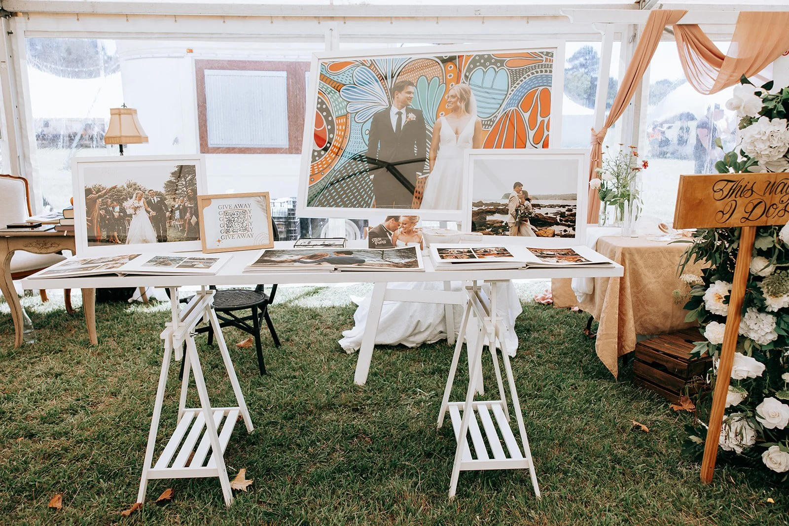 A wedding exhibition display featuring framed photos of couples, decorative floral arrangements, and signage on white tables in an outdoor tent setting.