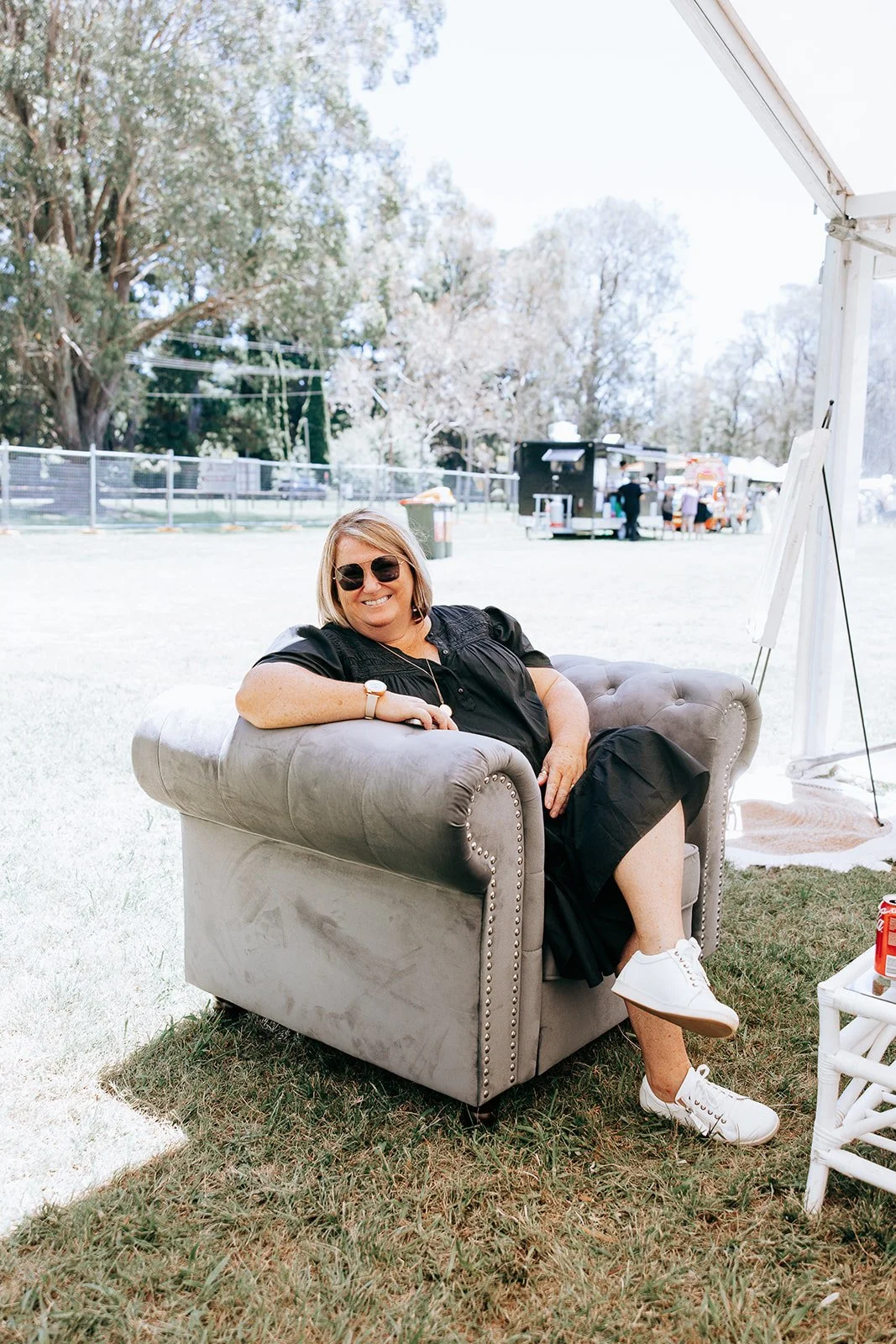 Person relaxing on a gray armchair outdoors at a park, wearing sunglasses and casual clothing. There's a white table with a drink can nearby, and food stands and trees in the background.