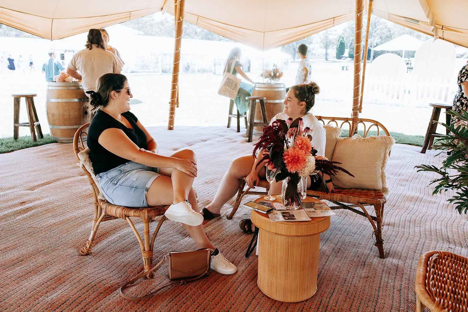 Two women sitting on wicker chairs under a tent, engaged in conversation. A wooden table with a floral arrangement and pamphlets is in front of them. There are other people in the background near wine barrels. The setting is outdoors on a sunny day.
