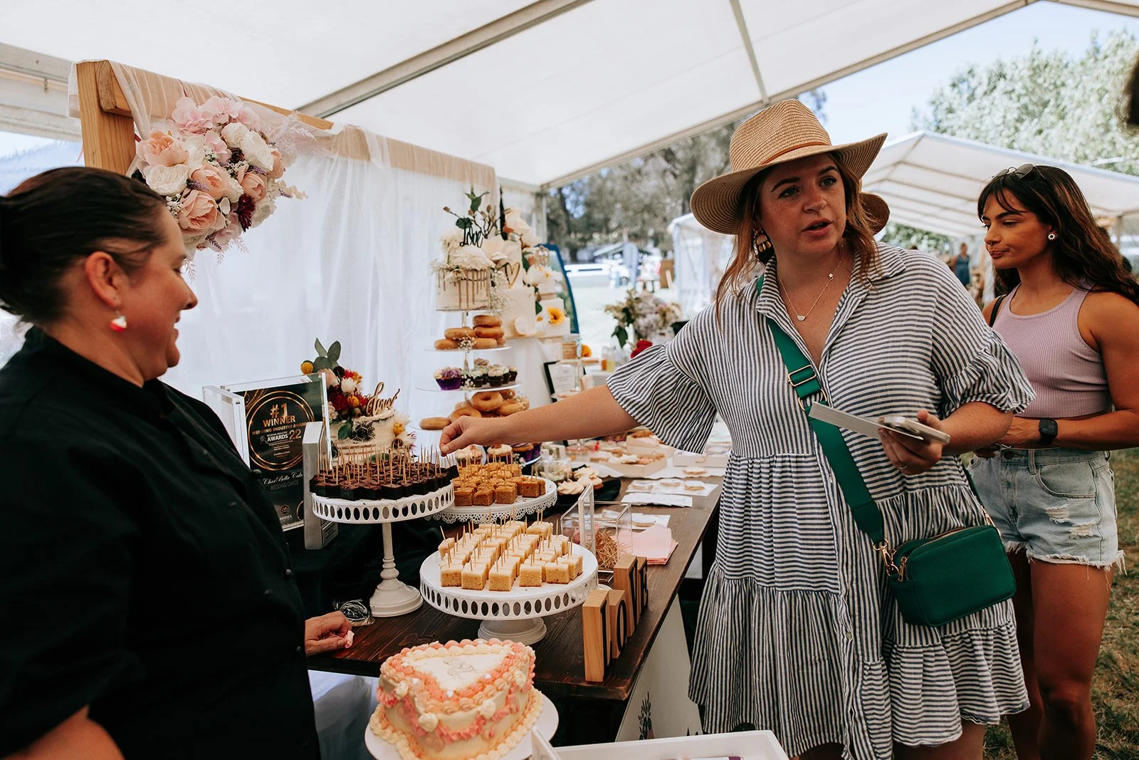 People at a bakery stall with various pastries and cakes on display under a tent.