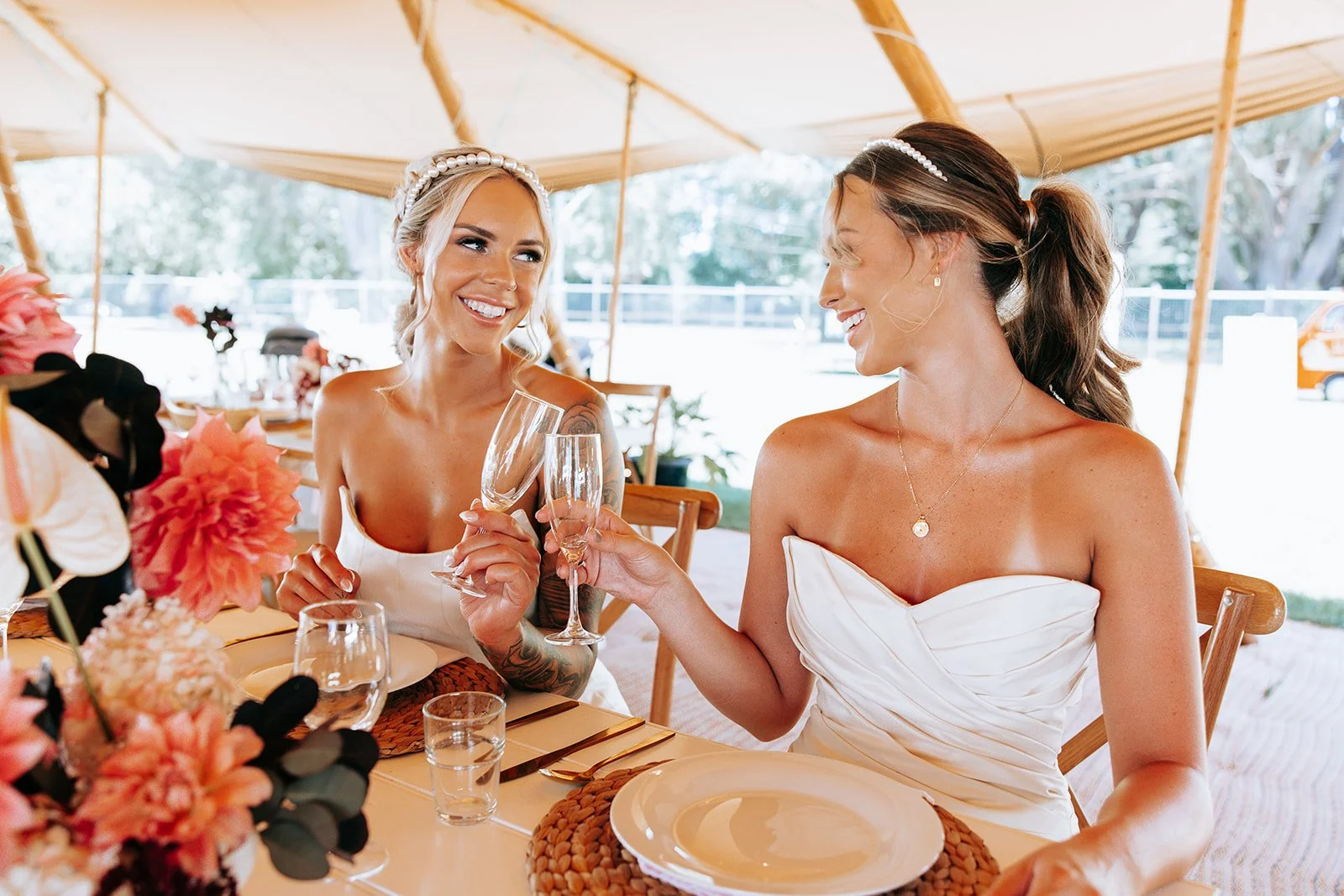 Two women in white dresses sitting at a table, smiling and clinking champagne glasses. The table is set with plates, glasses, and floral arrangements. They are under a tent.