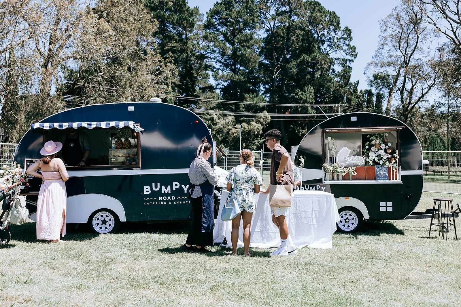 People interacting at a catering truck labeled "Bumpy Road Catering & Events" in a park setting, with trees and grass visible in the background. The scene suggests a casual outdoor event with attendees standing near a table set in front of the truck.