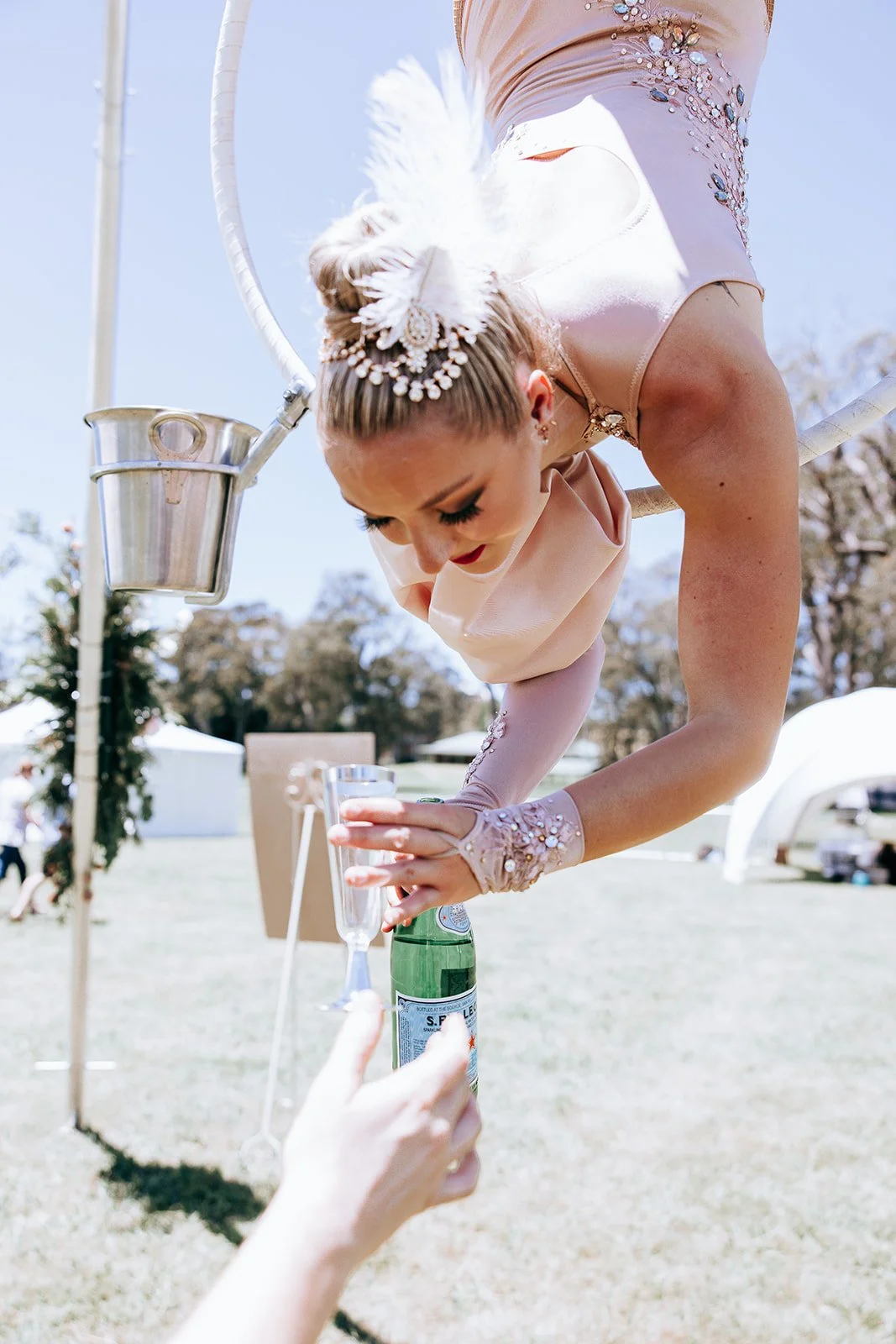 Acrobat suspended upside down pouring a drink into a glass outdoors.
