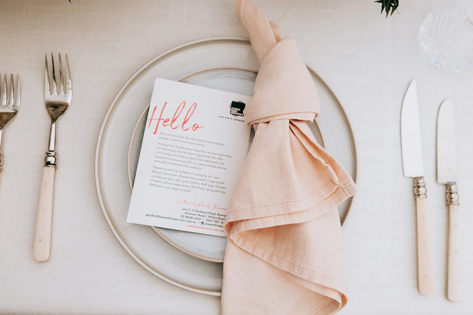Elegant table setting with a beige napkin, a hello note, and cutlery on a light plate.