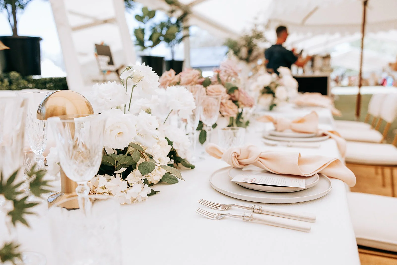 Elegant wedding table setting with white roses, pink napkins, crystal glasses, and gold accents under a tent.