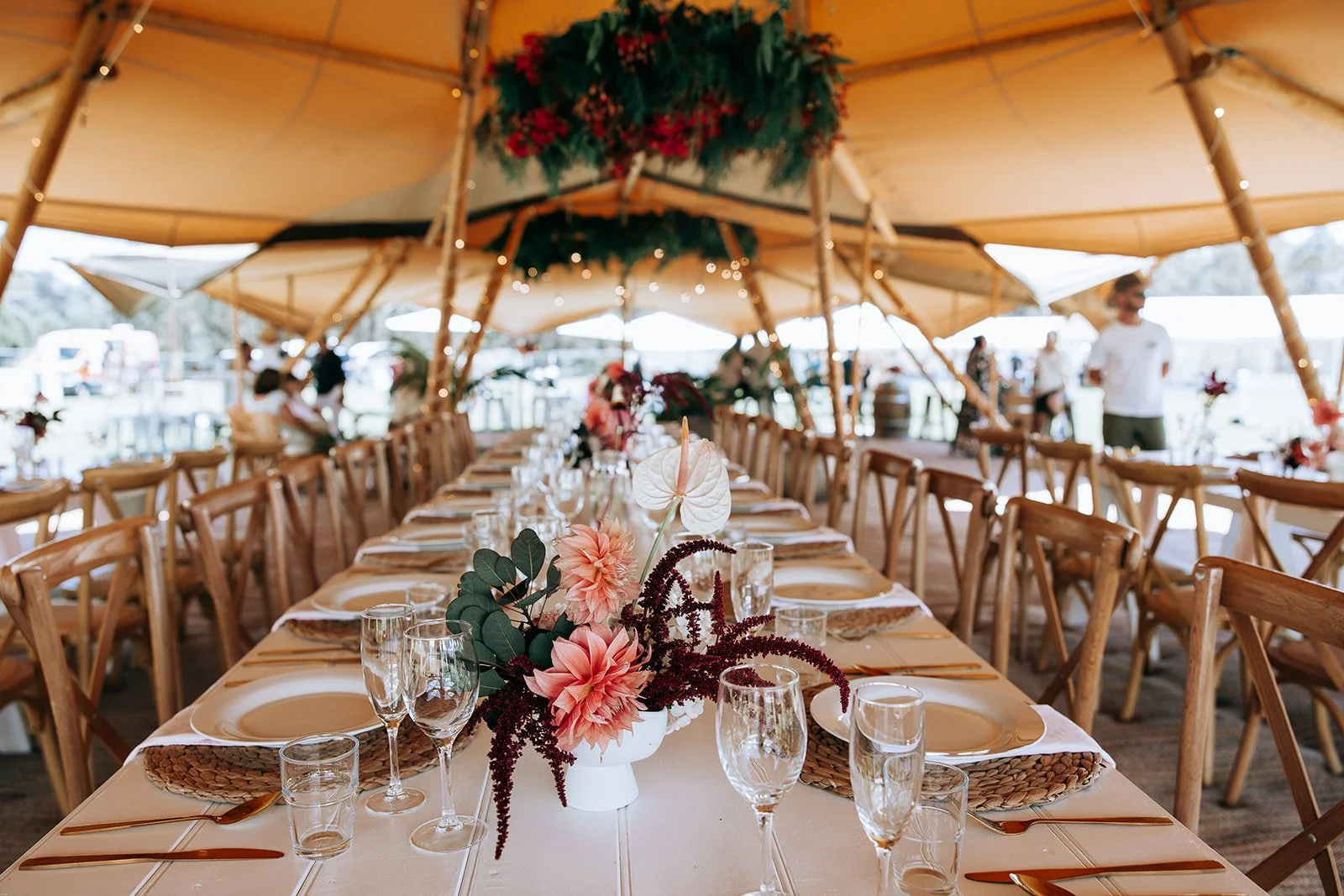 Elegant wedding reception in a tent with long tables, floral centerpieces, wooden chairs, plates, and glassware.