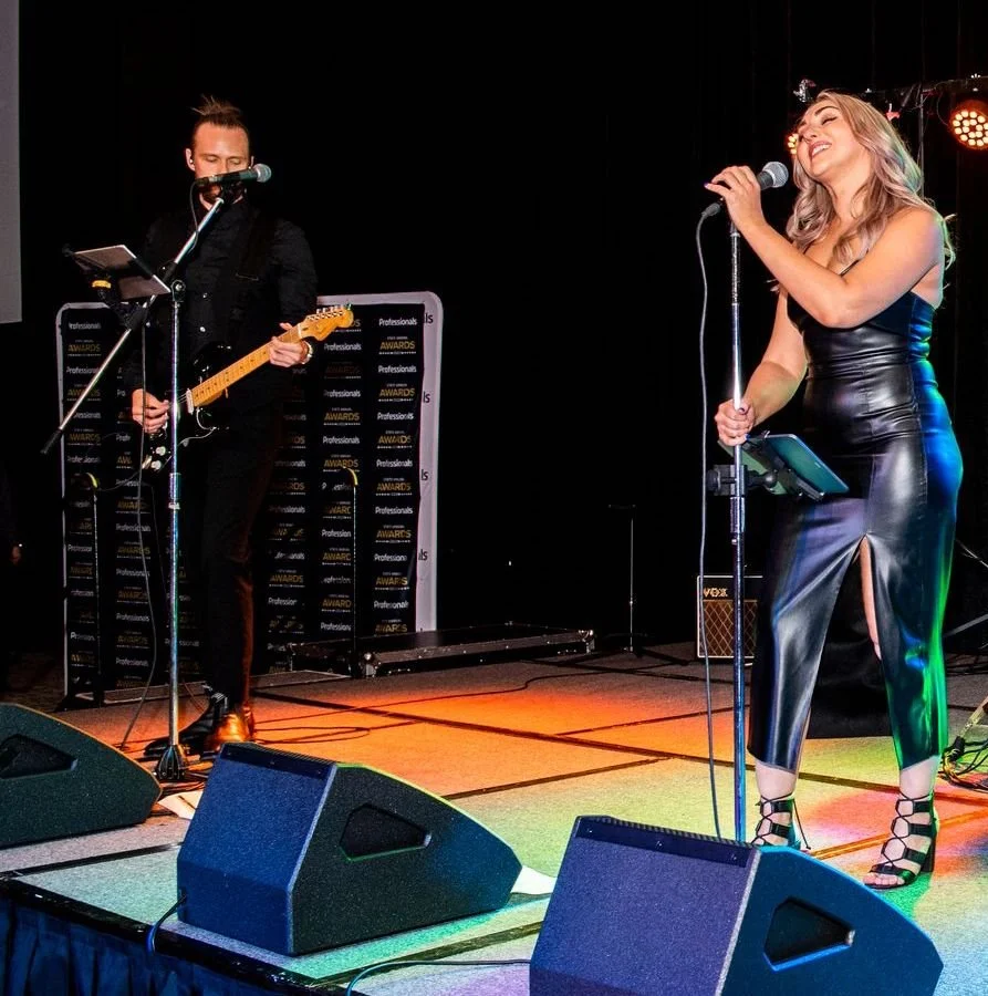 A singer performing on stage with a microphone, wearing a black leather dress and black high-heeled sandals, and a guitarist playing an electric guitar in the background, all under stage lights.