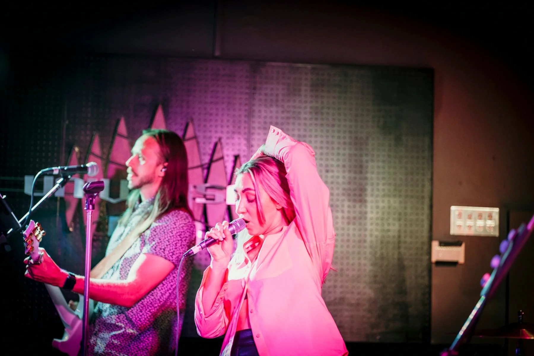 A woman singing into a microphone with her hand behind her head, and a man playing an electric guitar in the background, both on stage with colorful lighting.