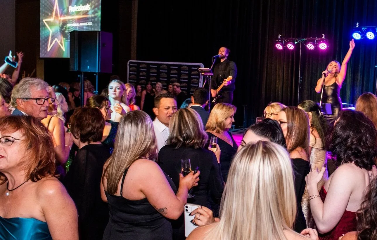 Crowd of people at a concert or party, with a live band on stage and colorful lighting.