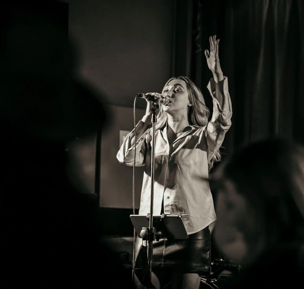 A woman singing into a microphone on stage, with one hand raised, in a black-and-white photograph.