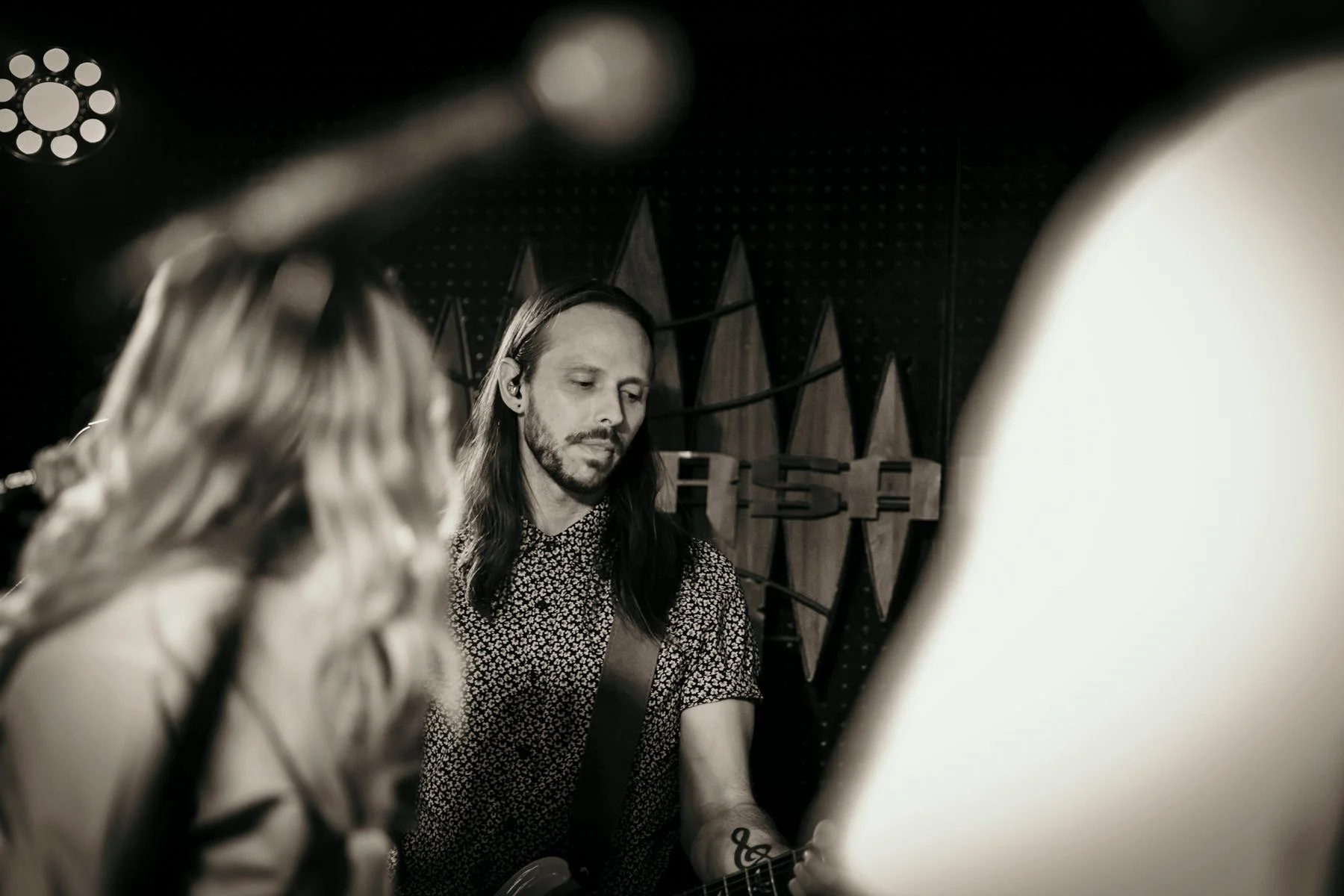 Black and white photo of a man with long hair playing an electric guitar, with a woman in the foreground and a perforated wall with geometric patterns in the background.