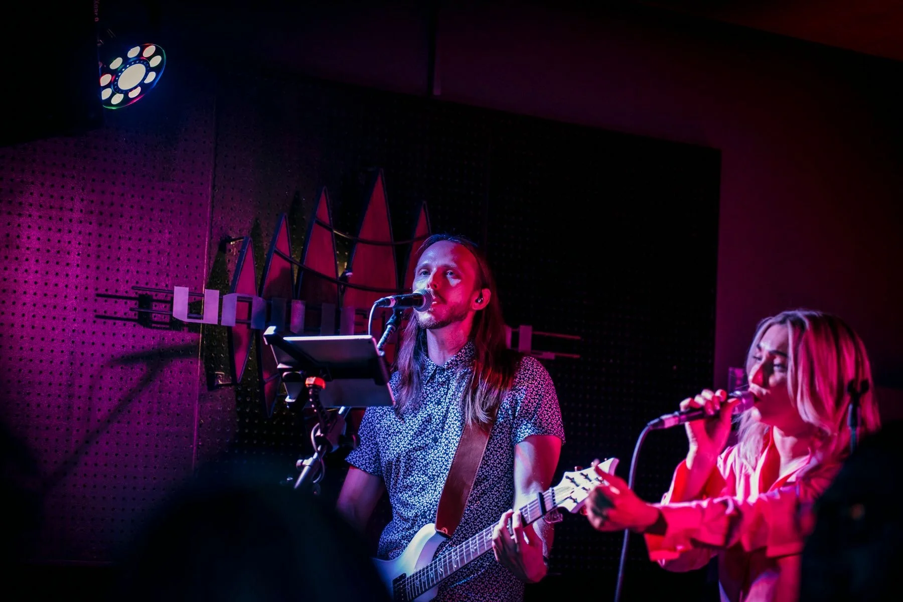 A woman playing guitar and singing into a microphone, with another woman singing into a microphone beside her, in a dimly lit stage with colored lights.
