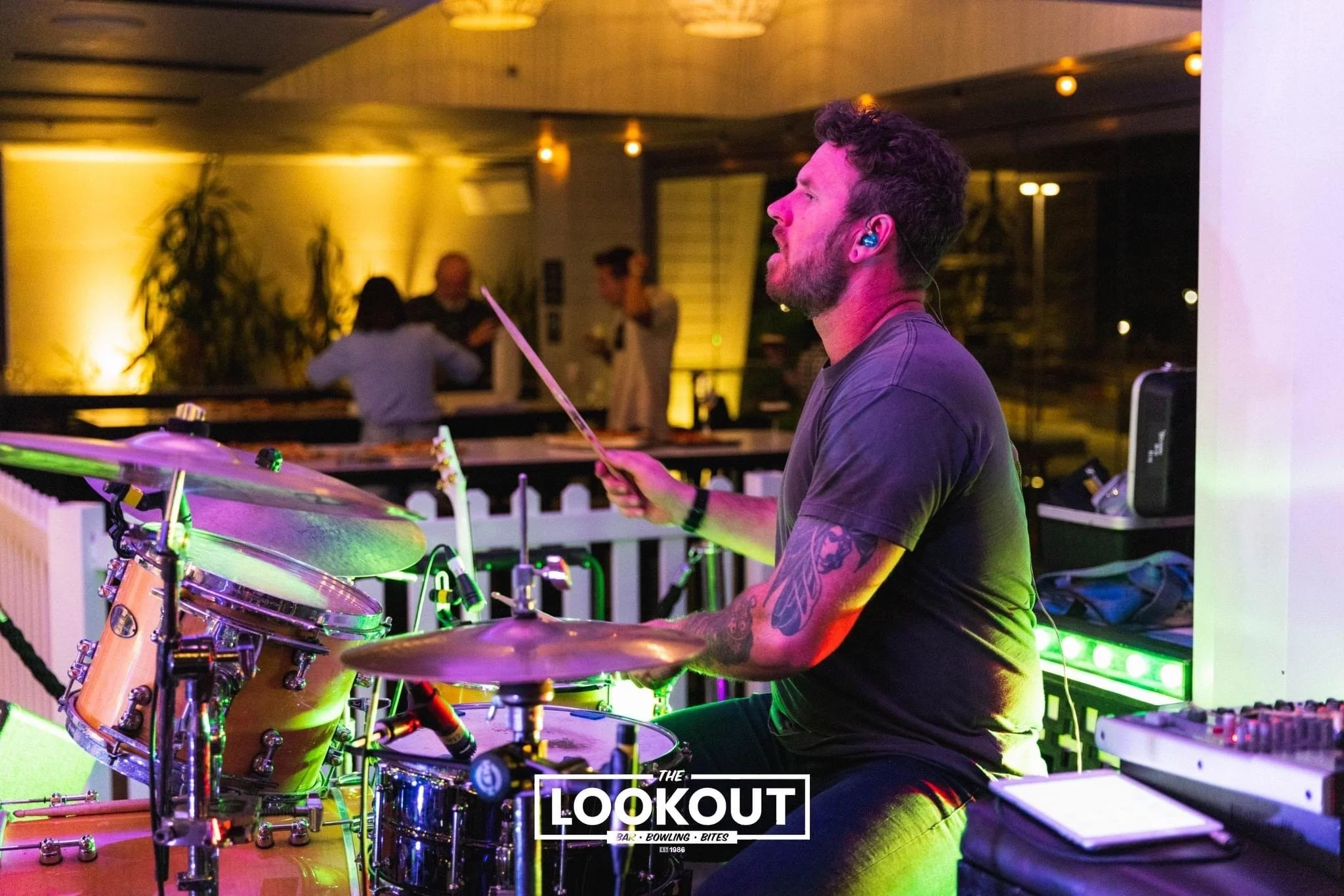 A man playing drums at a bar or club, with colorful lighting and people in the background, and the logo 'The Lookout Bar Bowling Bites' at the bottom.