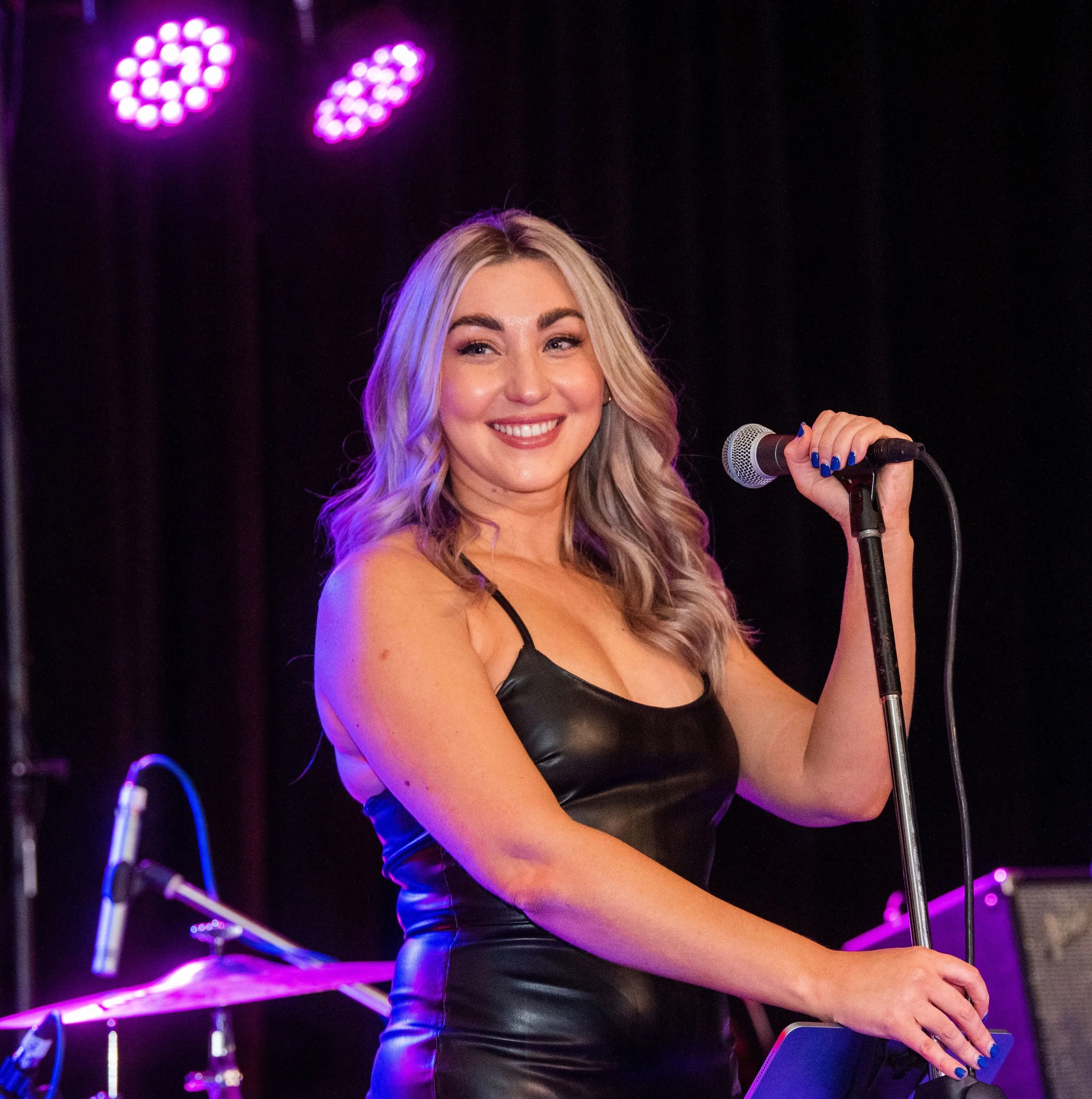 A woman with blonde hair wearing a black leather dress, holding a microphone on stage with purple lights in the background.
