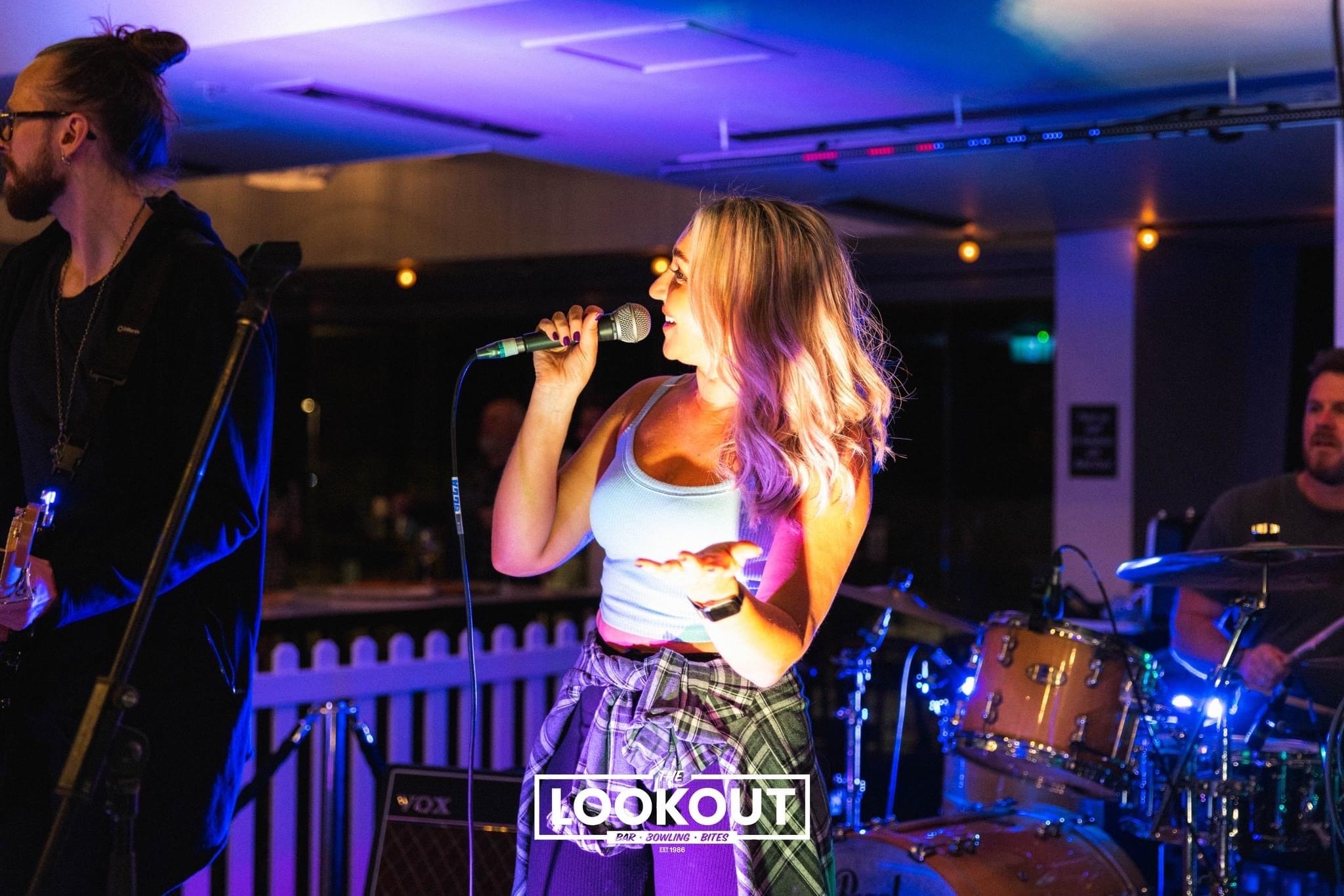 A woman with long blonde hair singing into a microphone during a live band performance, with drums in the background, at The Lookout Bar in Bowling Bites. She is wearing a white tank top and has a plaid shirt tied around her waist.