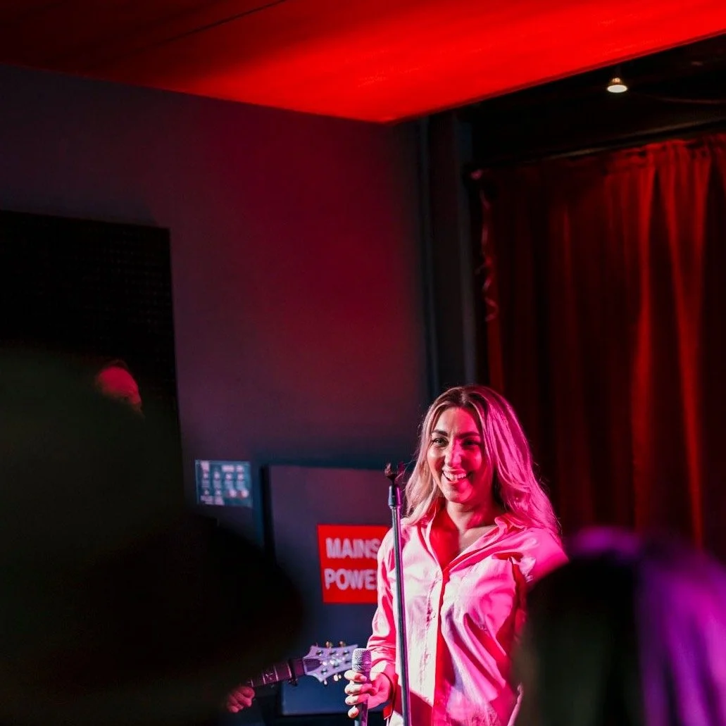 A woman with blonde hair smiling on stage, holding a microphone, with a guitar visible in the background, red and purple stage lighting, and a dark background with a sign.