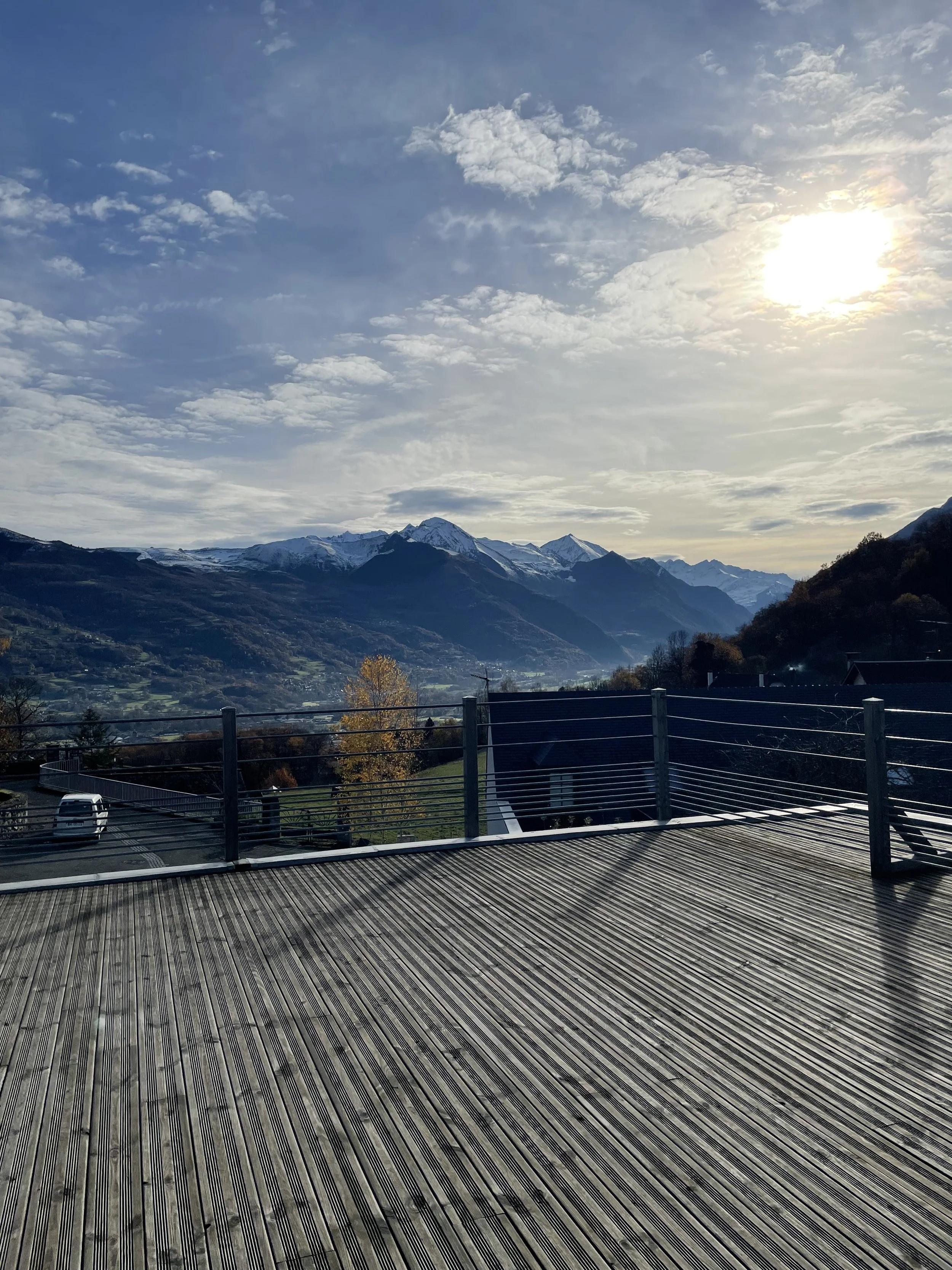 Vue d'un balcon en bois avec une vue sur des montagnes enneigées, un ciel partiellement nuageux et le soleil brille à l'horizon.
