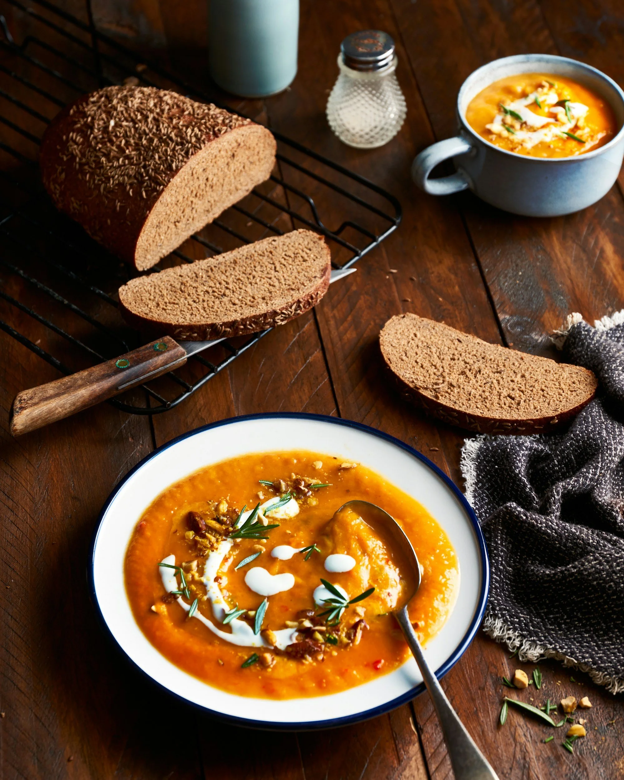 Soupe de potiron dans un bol blanc avec garnitures de neuro, crème, et herbes, accompagnée de tranches de pain complet sur une nappe en tissu.