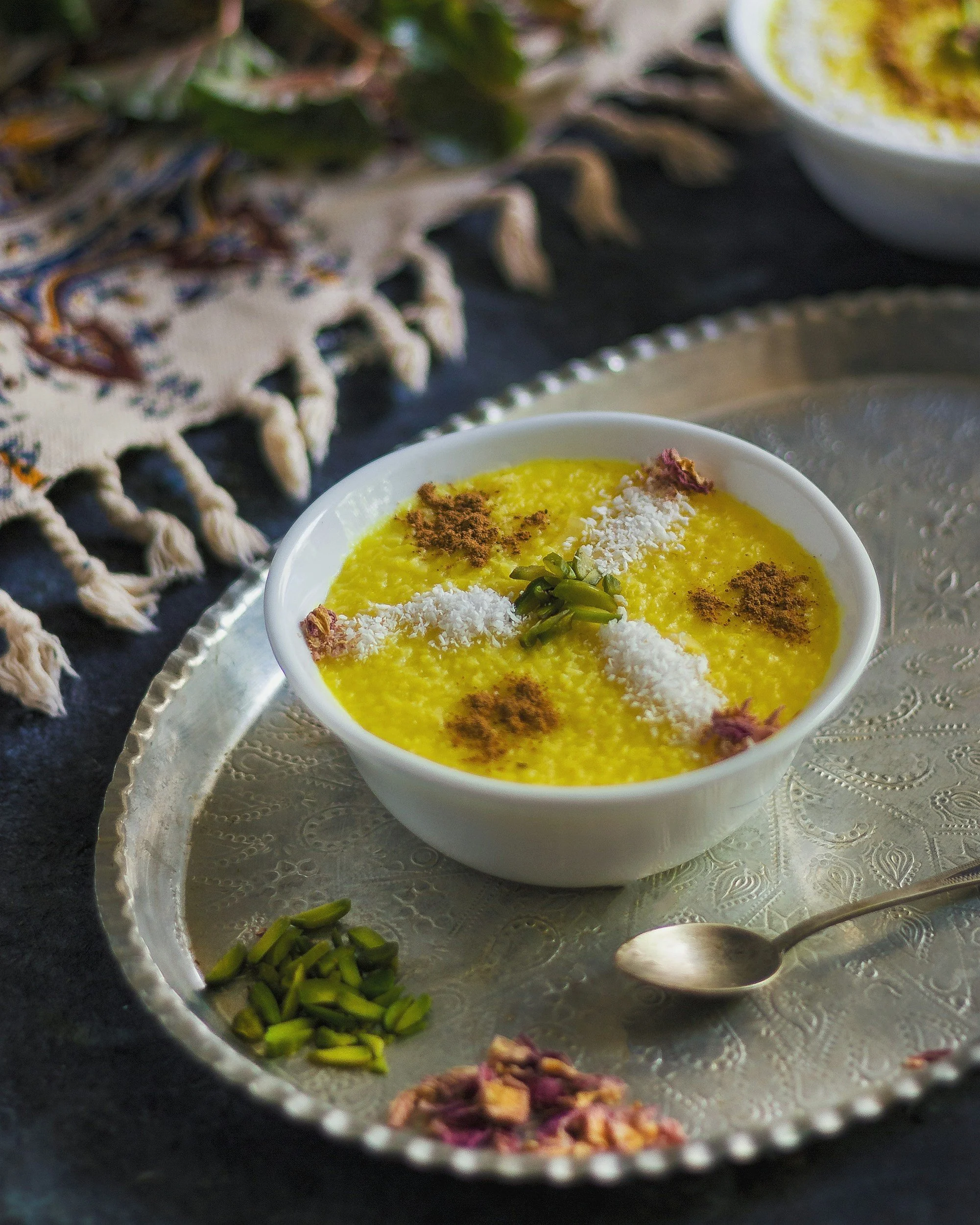 Un bol de kheer, un dessert indien au riz, parsemé de noix de coco râpée, de noix, de feuilles de cardamome et de poudre de cannelle, placé sur un plateau en métal argenté avec d'autres plats et une nappe brodée en arrière-plan.