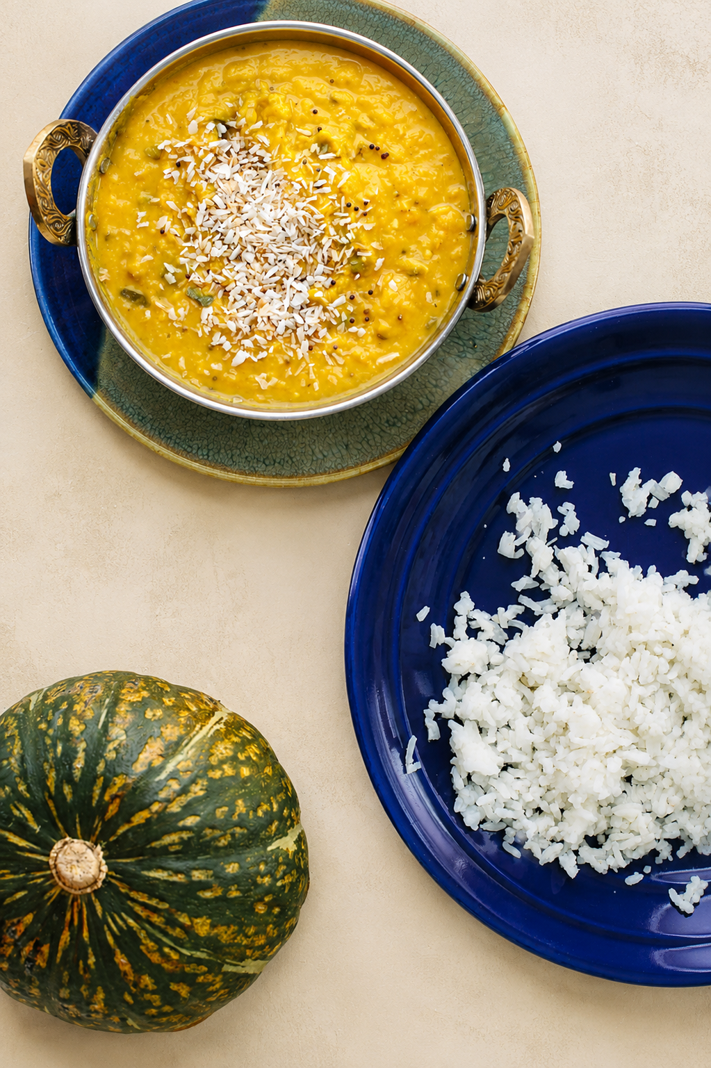 Une courge verte et jaune, un plat de riz blanc haché sur une assiette bleue, et un bol de dhal jaune garni de noix de coco râpée, sur une surface claire.