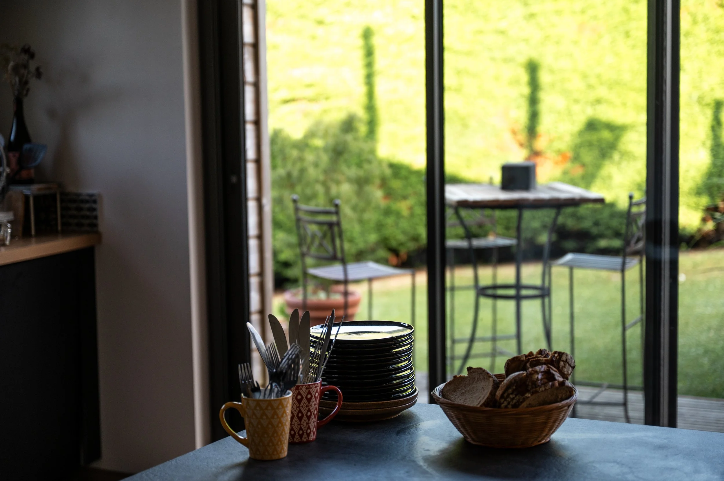 Cuisine intérieure avec assiettes empilées, couverts dans deux mugs colorés, un panier de pain sur la table, et vue sur un patio extérieur avec table et chaises autour d’un jardin vert.