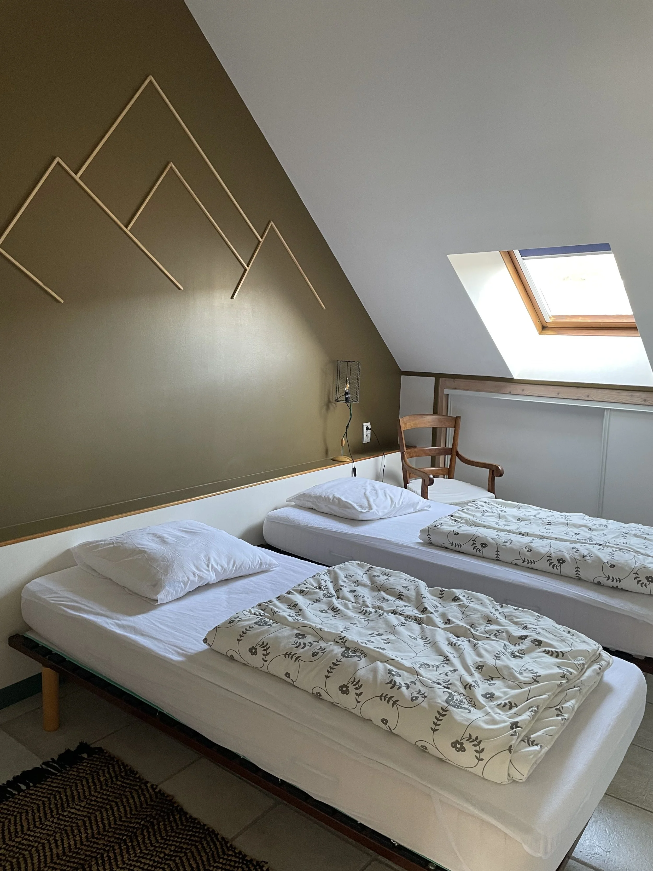 Chambre avec deux lits simples, une chaise en bois, un toit en pente avec une fenêtre de toit, une décoration murale abstraite, et une lampe murale à côté des lits.