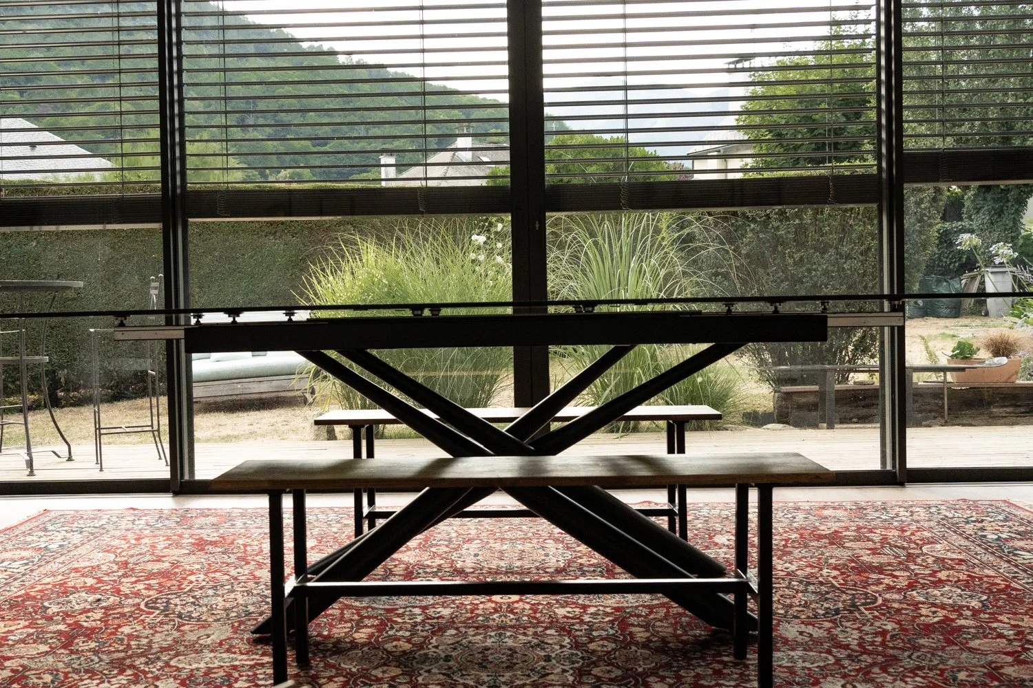 Intérieur d'une salle avec une table de ping-pong, un tapis orangé à motifs, et un grand mur vitré donnant sur un jardin avec des plantes et une terrasse en bois.