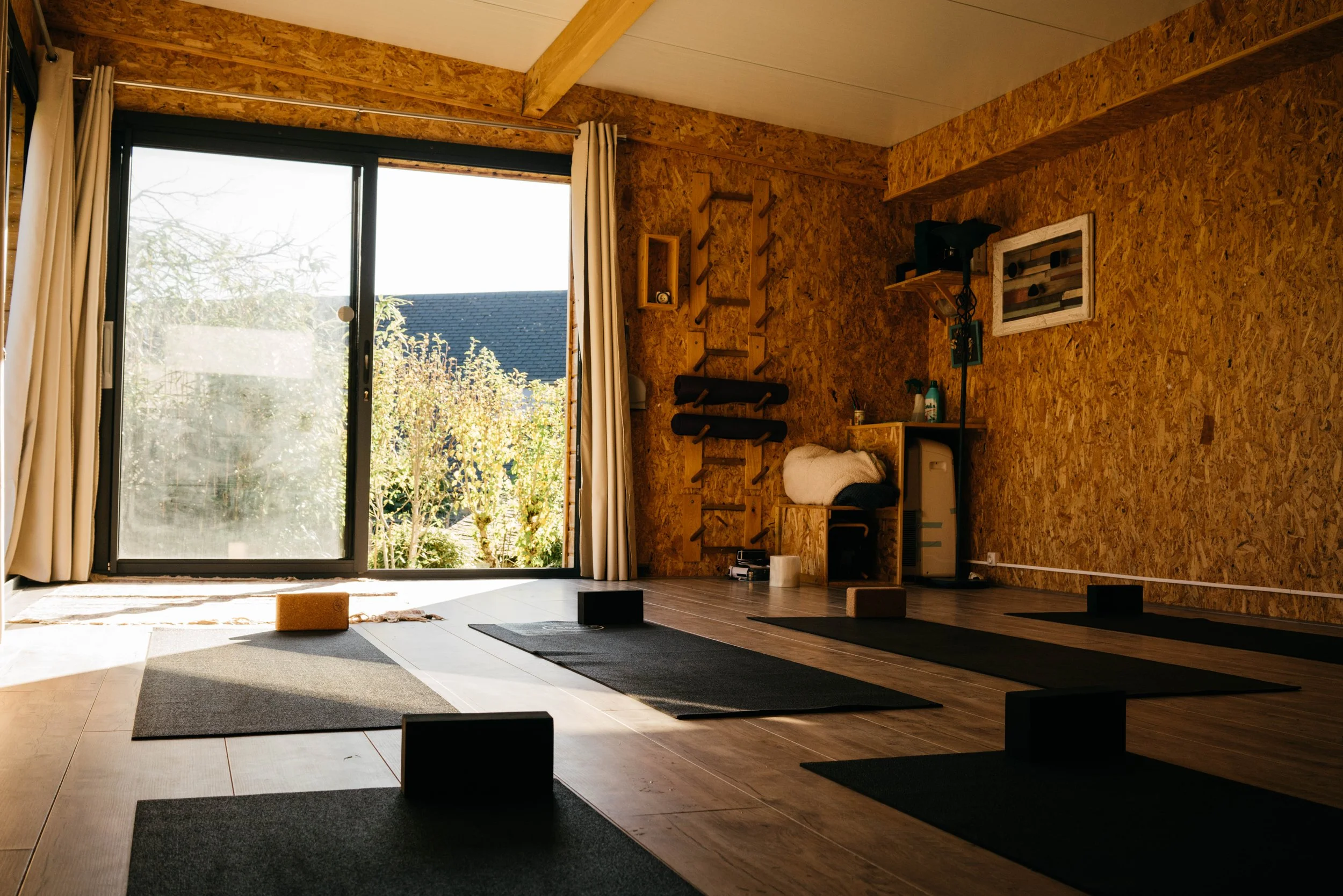 Salle de yoga avec tapis, blocs de yoga et fenêtres donnant sur un jardin.