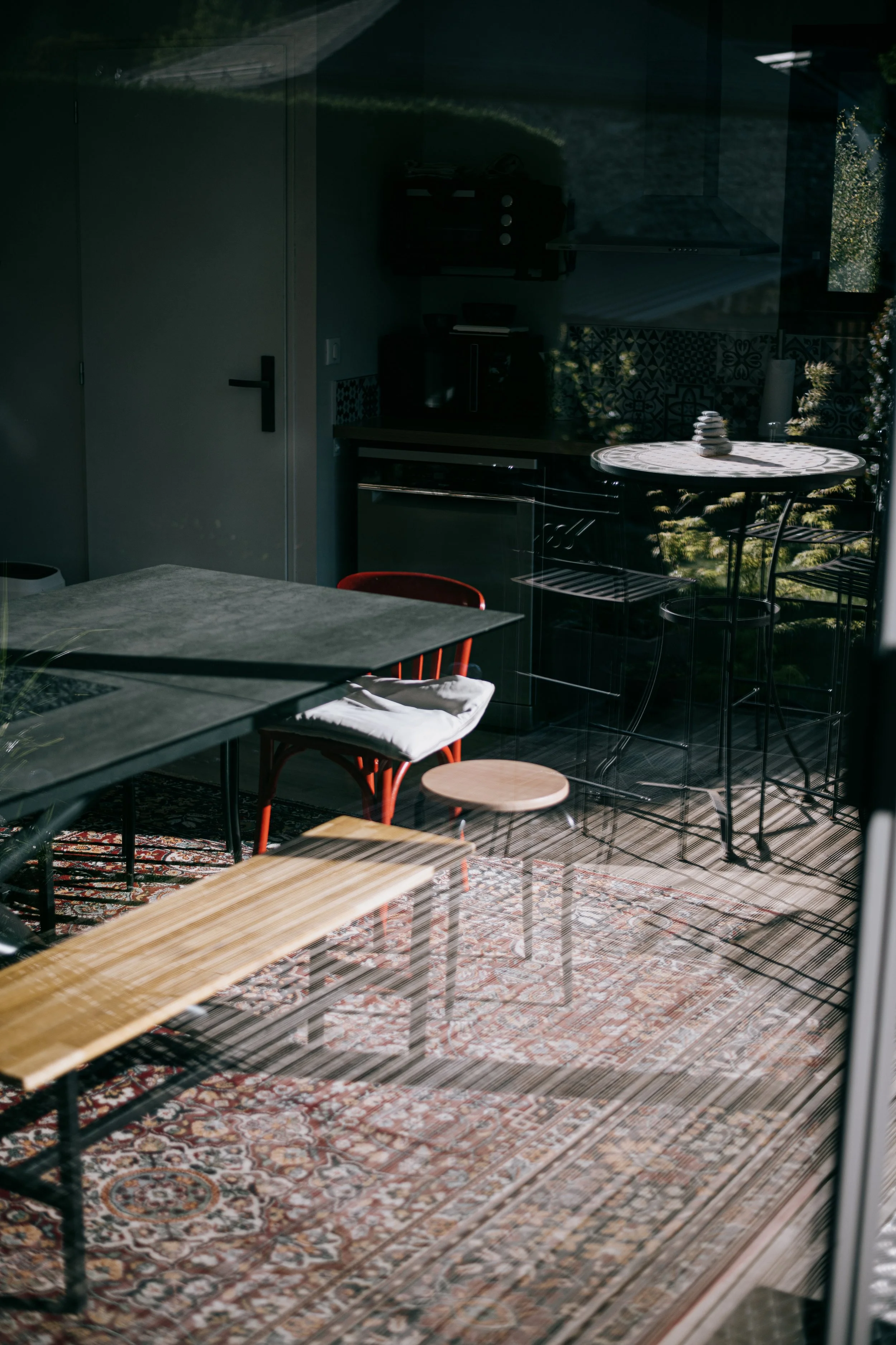 Une cuisine intérieure vue à travers une fenêtre avec des meubles en bois, une table ronde avec des pierres, un tapis, un chariot haut, et des chaises, avec la lumière du soleil créant des ombres.