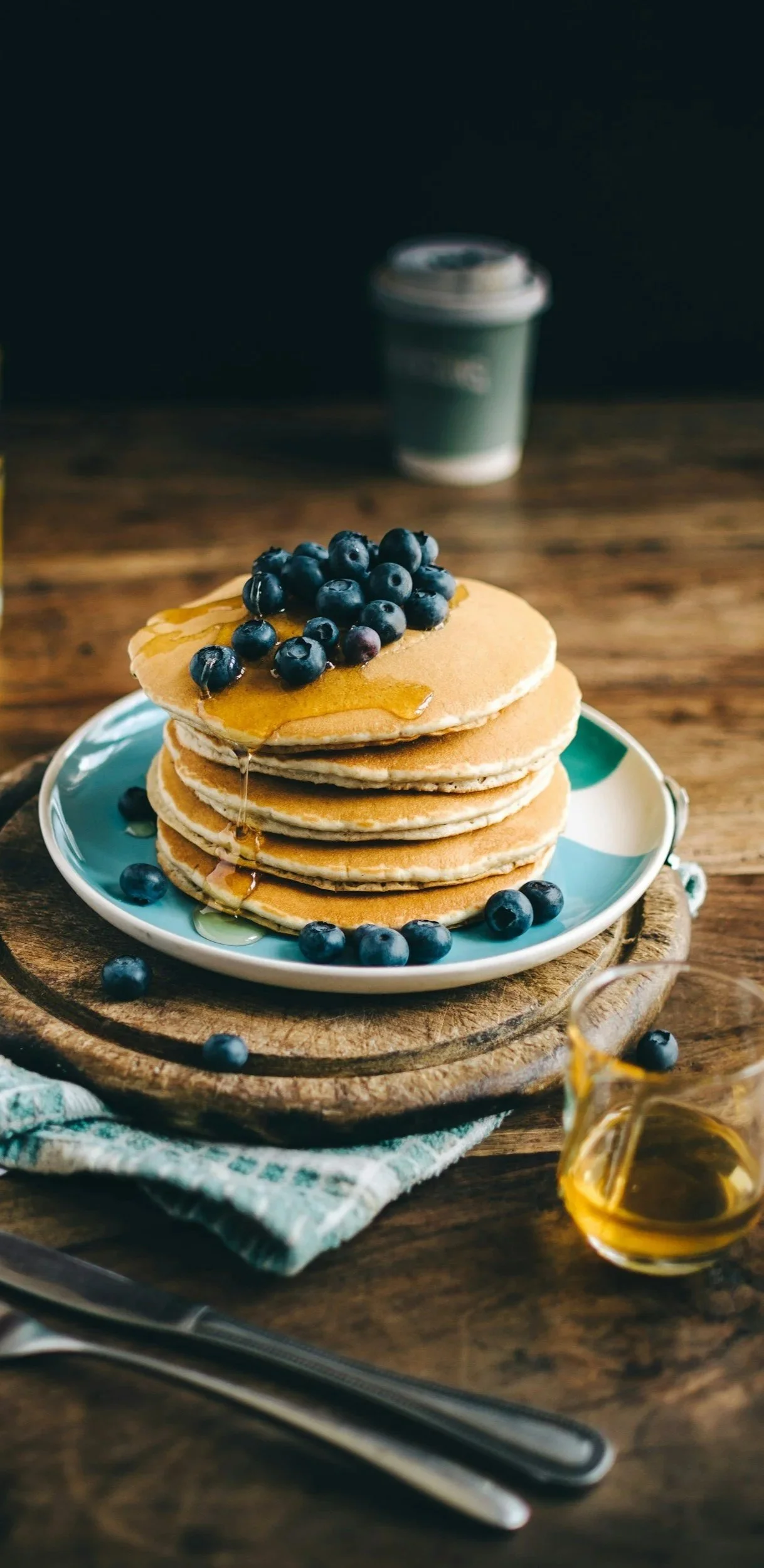 Une pile de pancakes garnis de myrtilles, de miel, servie sur une assiette colorée, avec une tasse à café en arrière-plan. L'ensemble repose sur une table en bois.