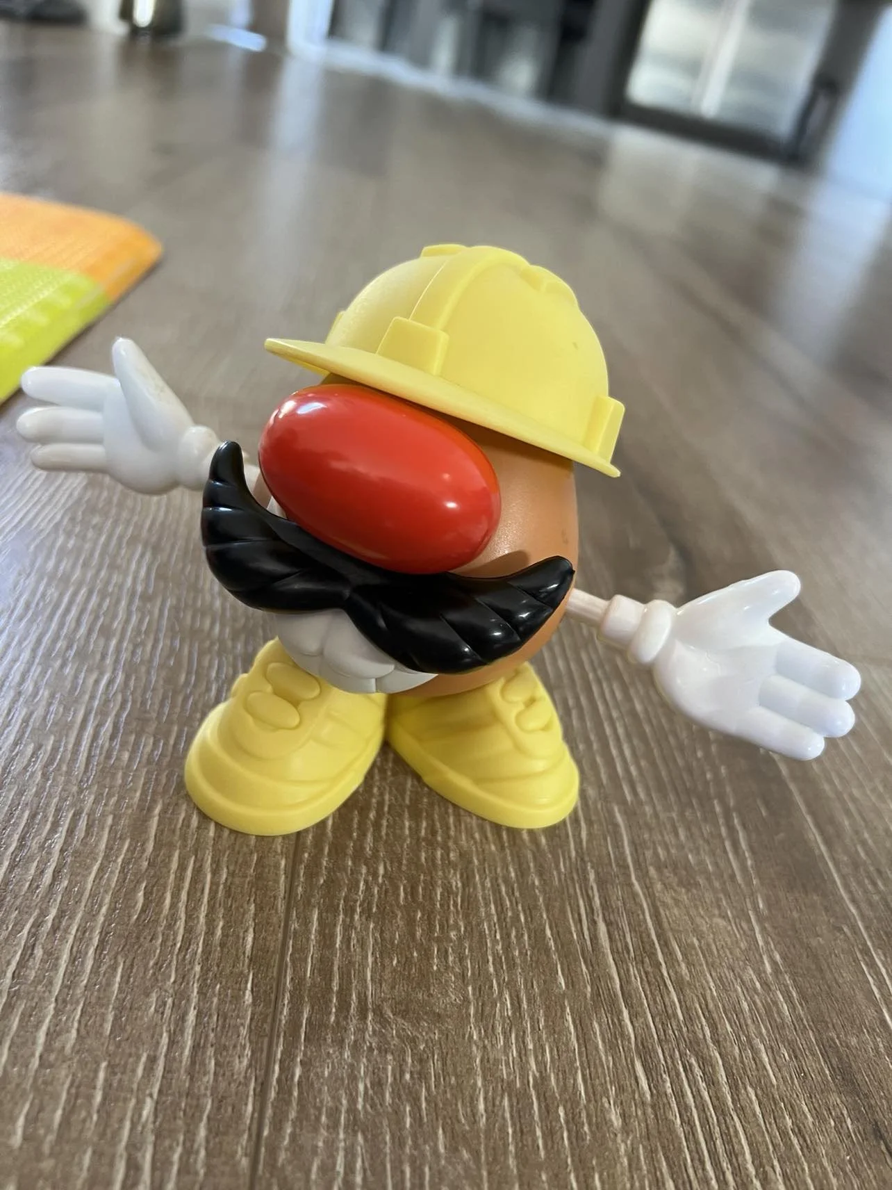 Toy figure resembling Mario, wearing a yellow hard hat and yellow shoes, with a red nose, black mustache, white gloves, and standing on a wooden surface.