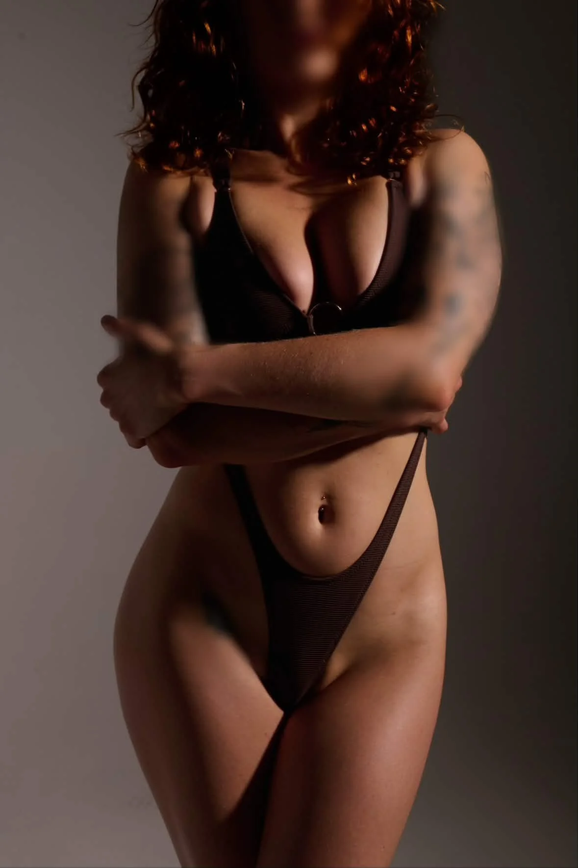 A woman with curly red hair wearing black lingerie, standing with her arms crossed over her chest, in a studio setting with soft lighting.