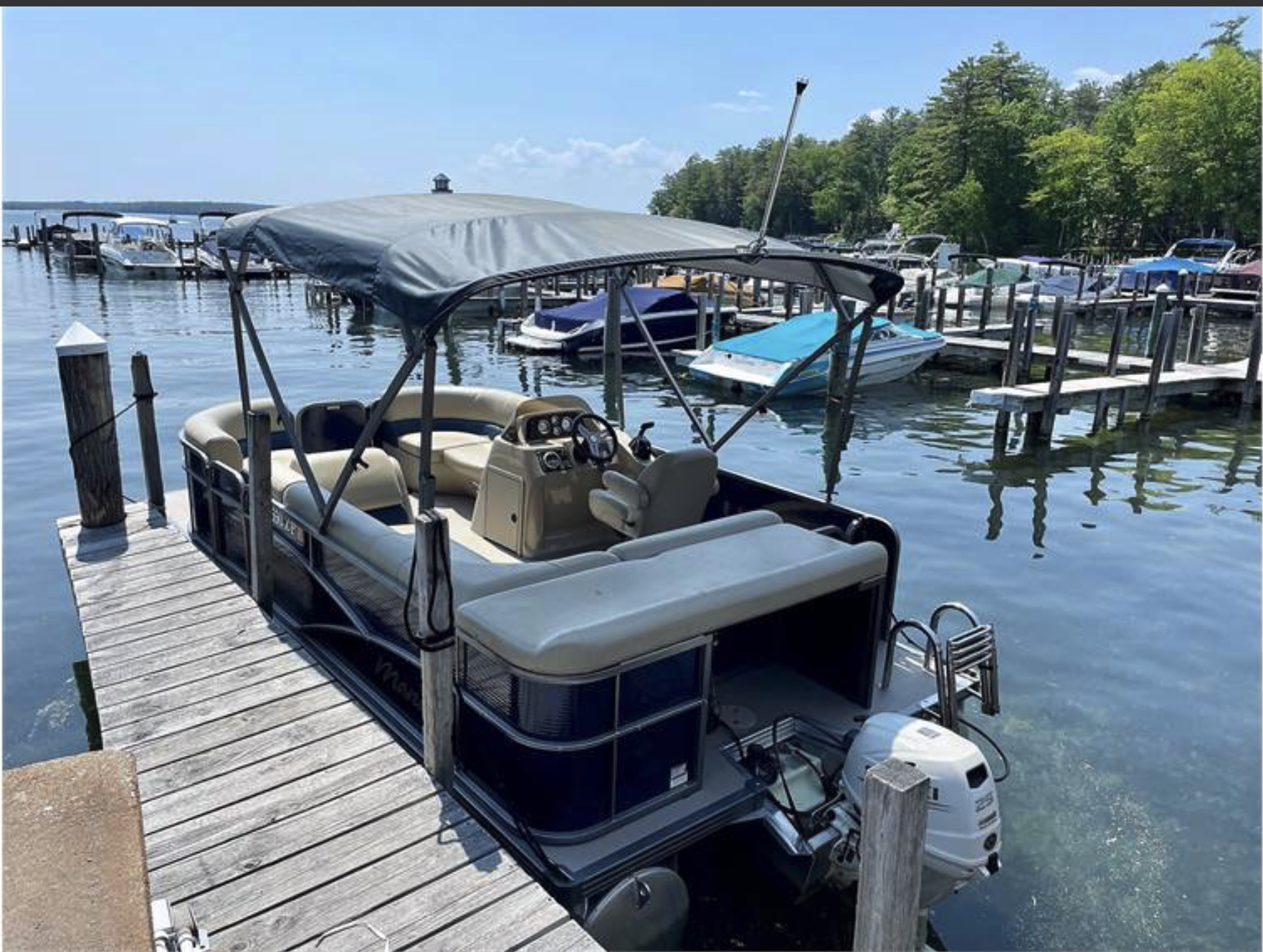 A pontoon boat docked at a wooden pier on a lake, with other boats in the background, and a bright orange sale tag that reads 'Weekly Special $2100'.