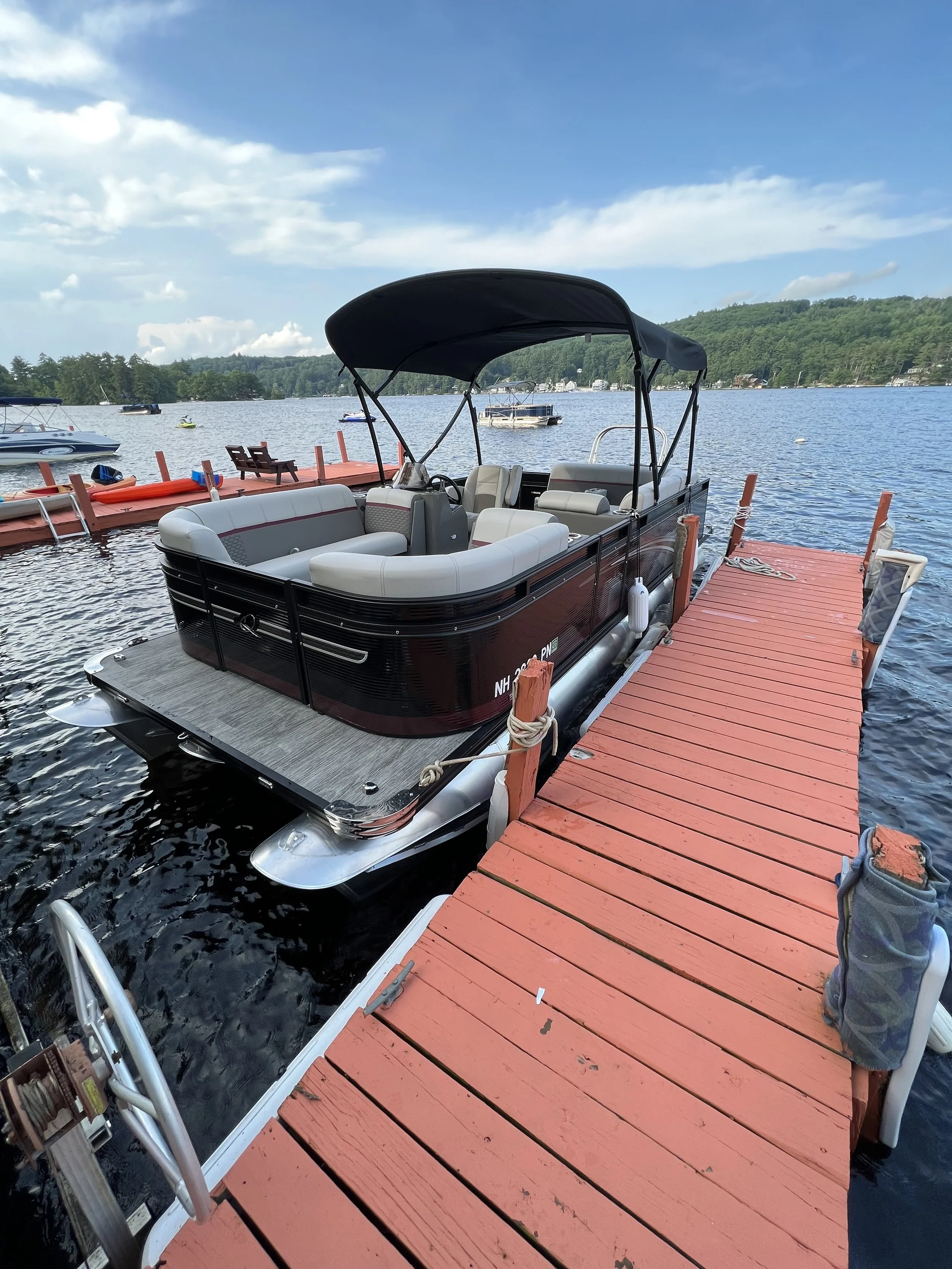 A black and gray pontoon boat docked at a red wooden dock on a calm lake with boat rentals and green forest in the background. A promotional graphic indicating a weekly special price of $2622.00.