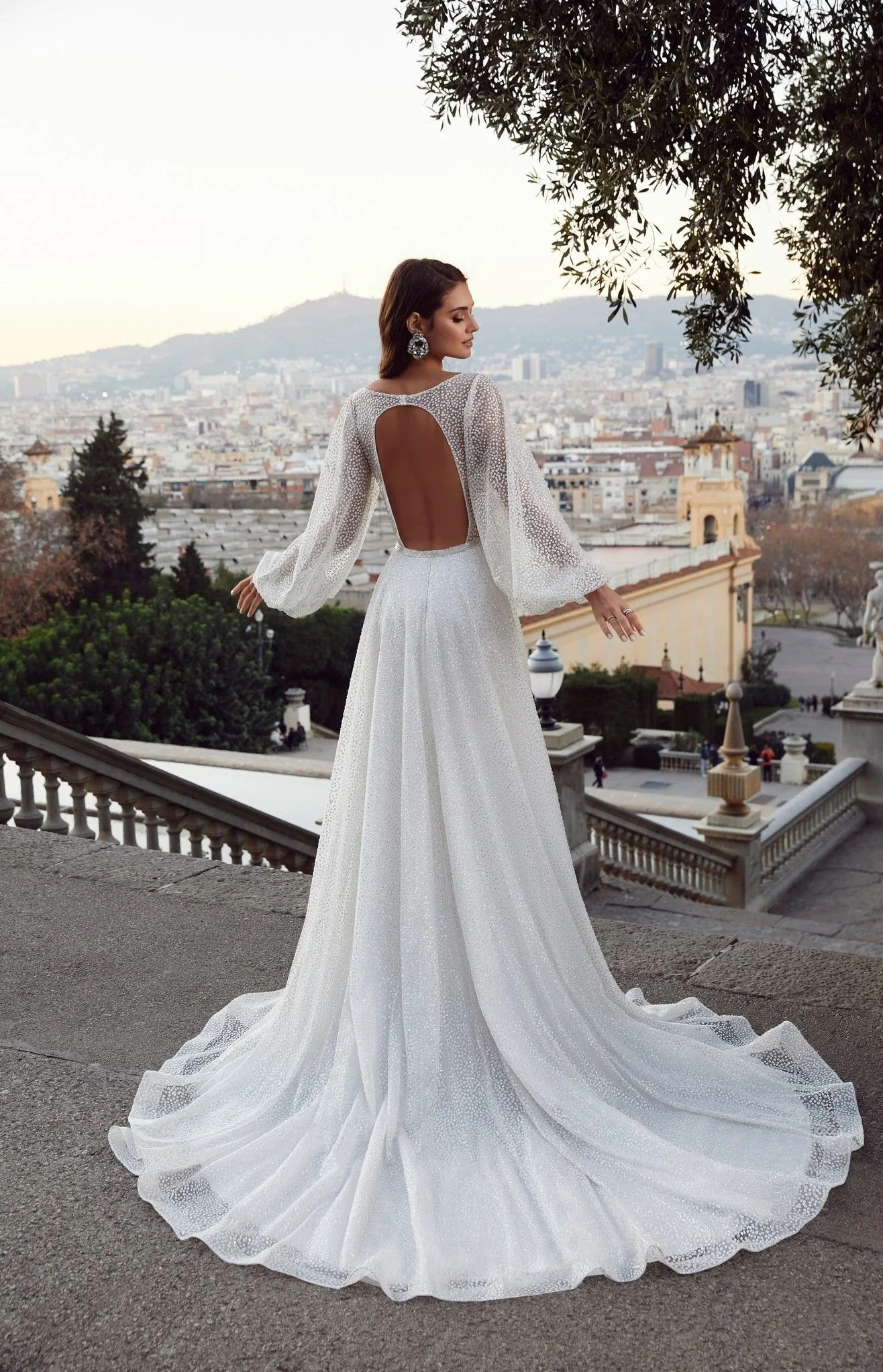 Woman in long white gown with open back standing outdoors with cityscape view.