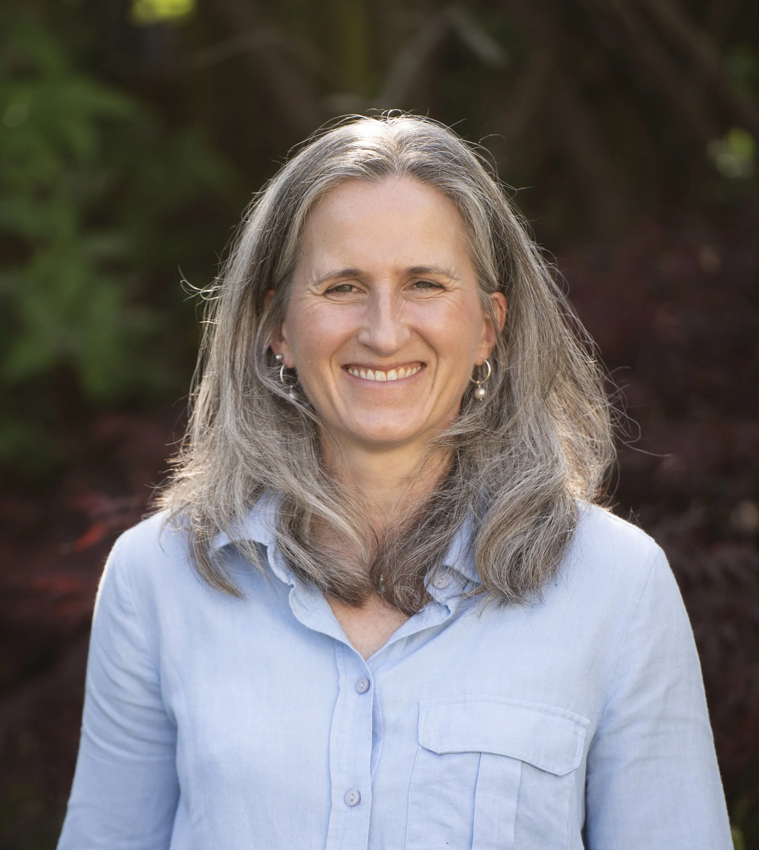 Close-up photo of a woman with long gray hair smiling, wearing pearl earrings and a light blue collared shirt, outdoors with blurred greenery in the background.