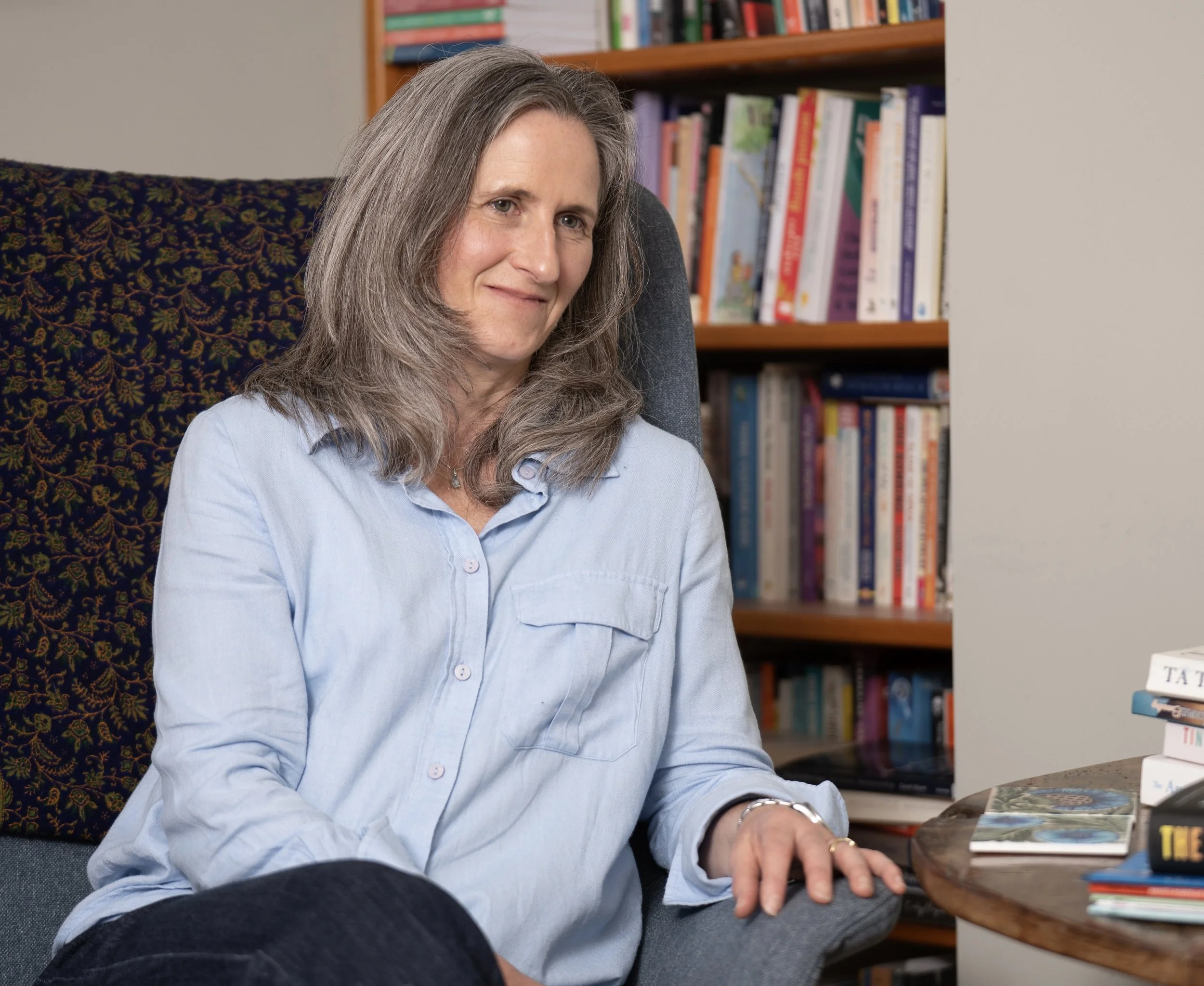 A woman with gray hair wearing a light blue shirt sitting in a room filled with books, looking at the camera with a slight smile.